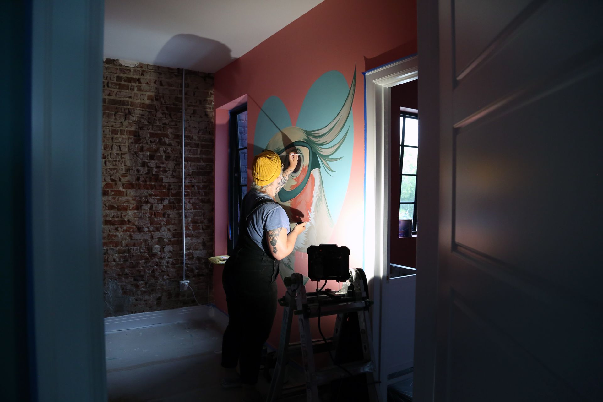 Person painting a colorful mural on a wall in a dim room with exposed brick and pink walls