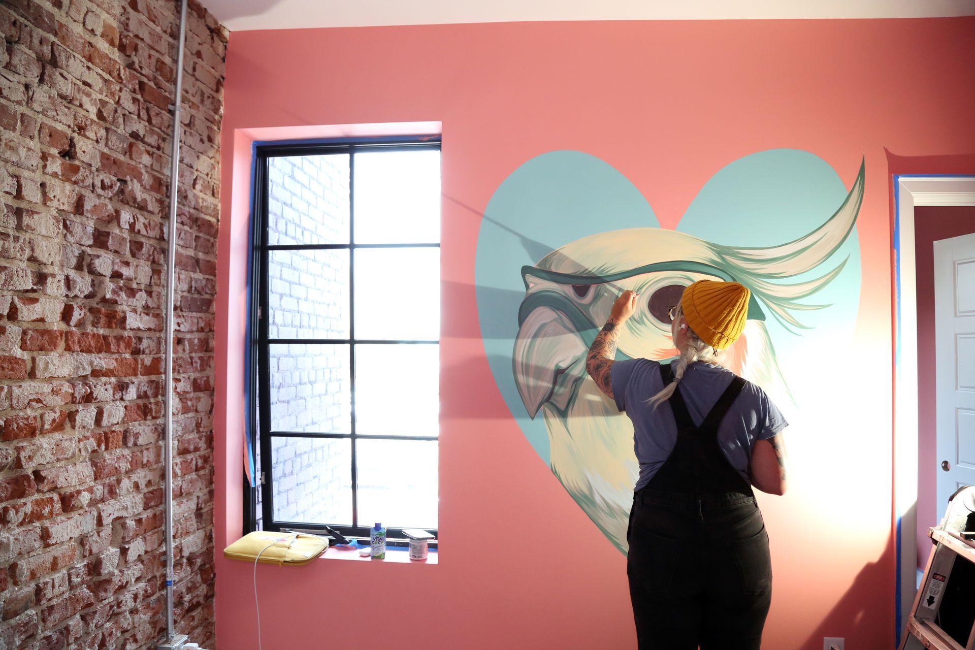 Person painting a large heart mural on a pink wall in a bright room