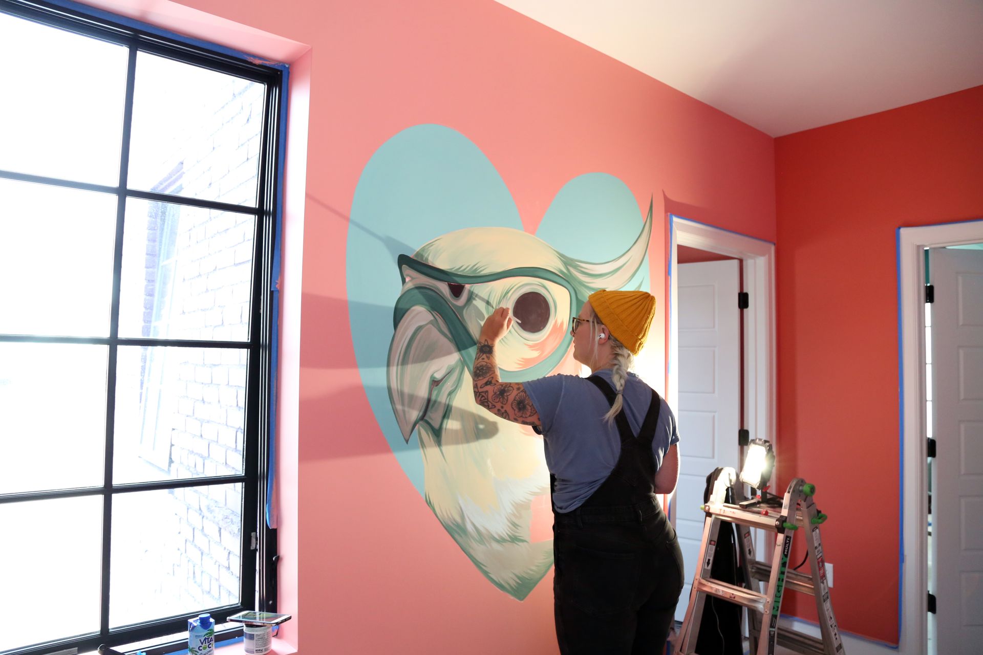 Person painting a mural of a bird on a pink wall in a room with large windows
