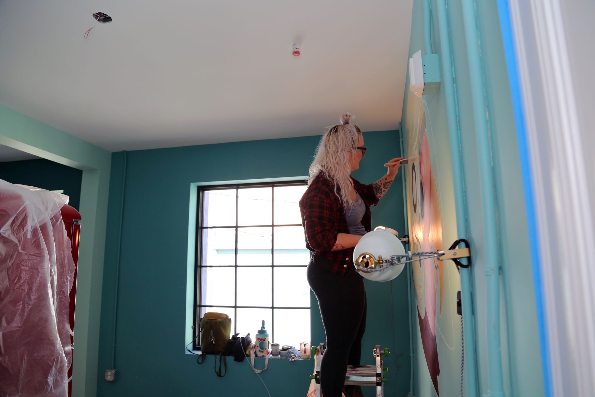 Person standing on a ladder painting a turquoise wall in a bright room