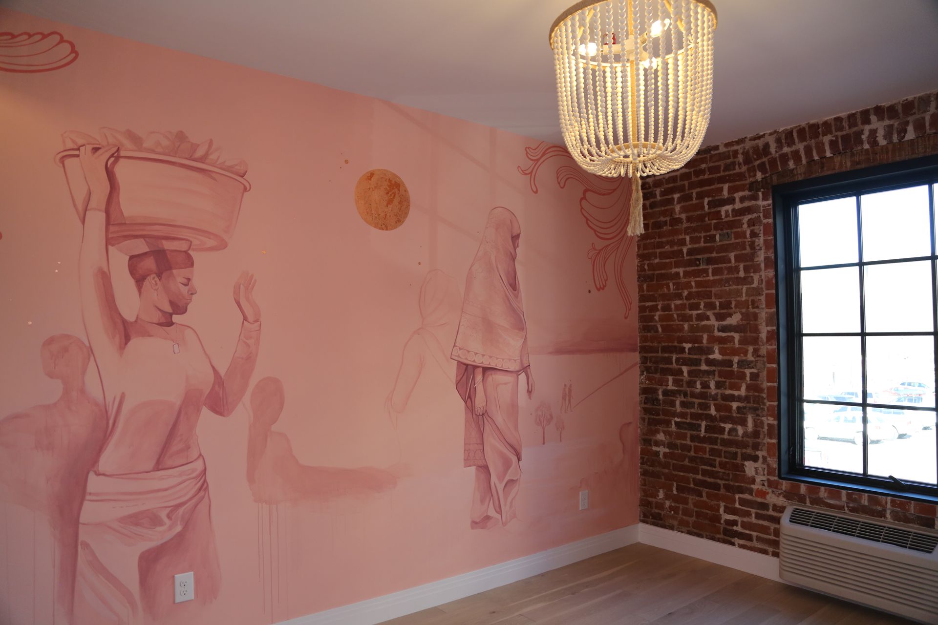 Pink muraled room with exposed brick wall, large window, and hanging crystal chandelier.