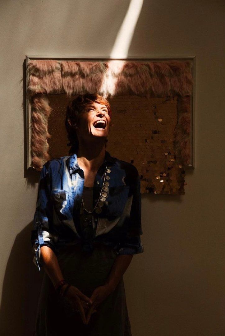 Woman in a dark dress, lit by a beam of light, smiling beneath a textured wall hanging.