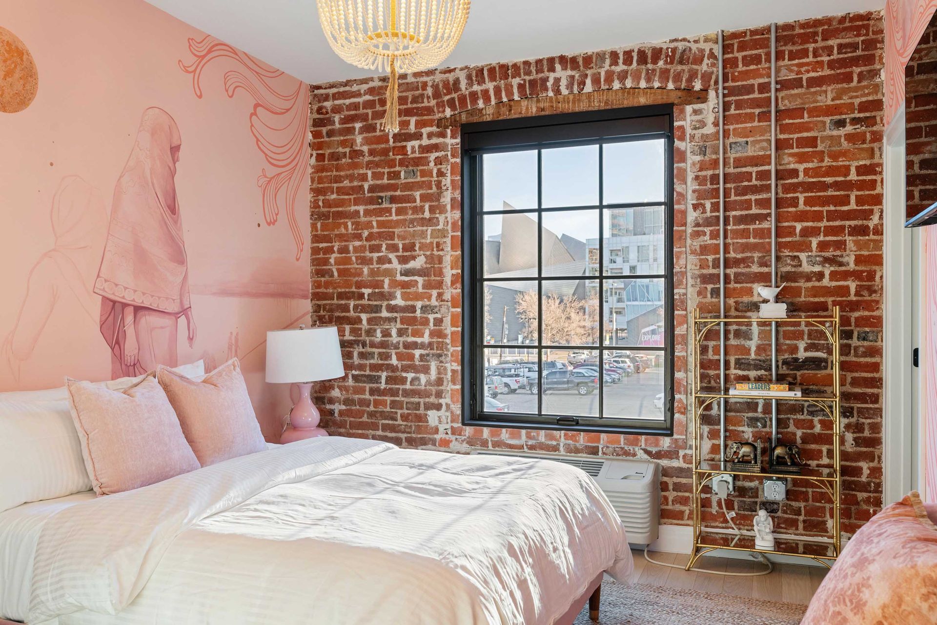 Bright bedroom with brick wall, pink accent wall, large window, and white bed with a pink throw