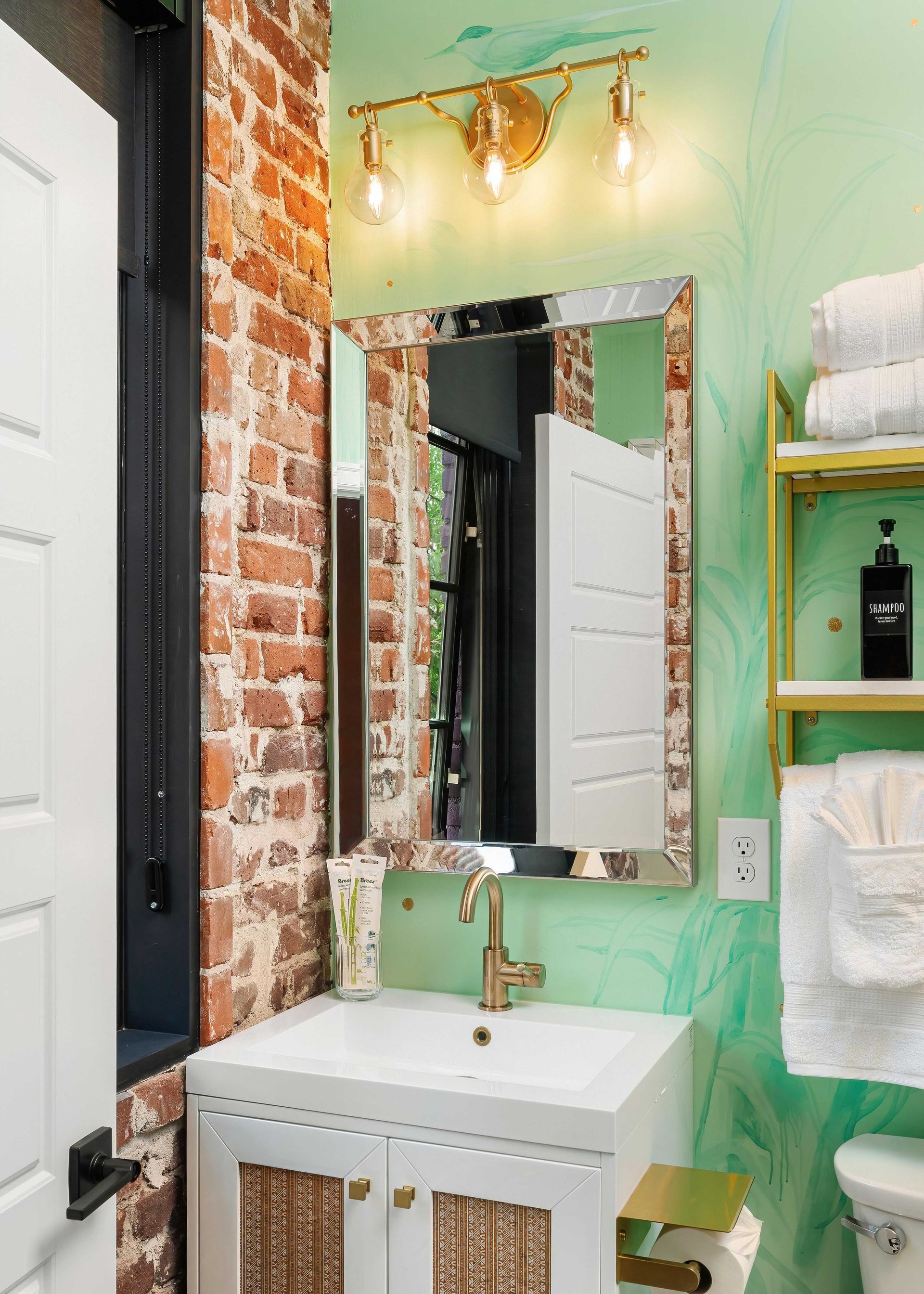 Small bathroom with mint-green walls, exposed brick, white sink, mirror, and towel shelves.