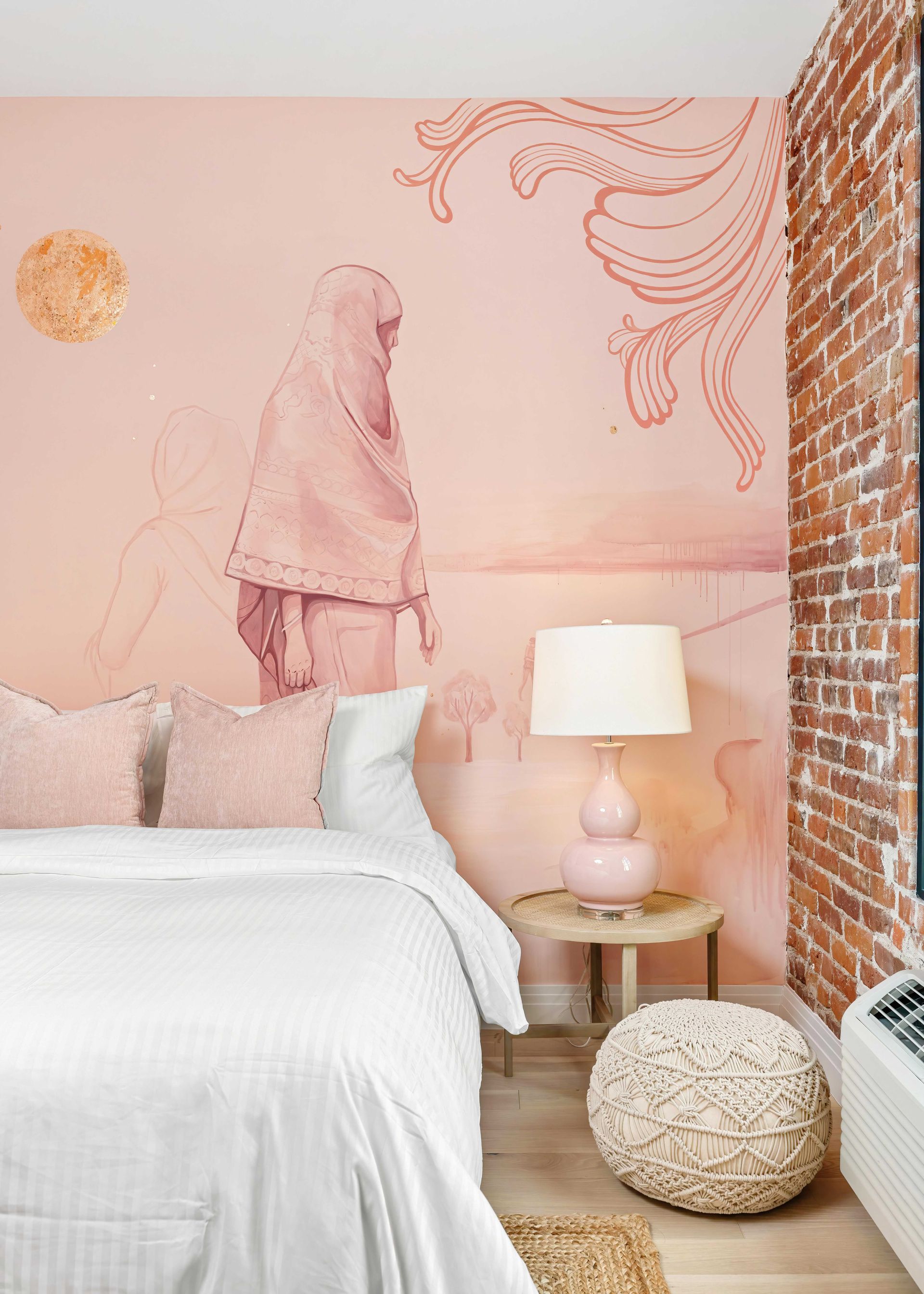 Pink bedroom with mural wall, white bed, bedside lamp, and woven pouf beside a brick accent wall