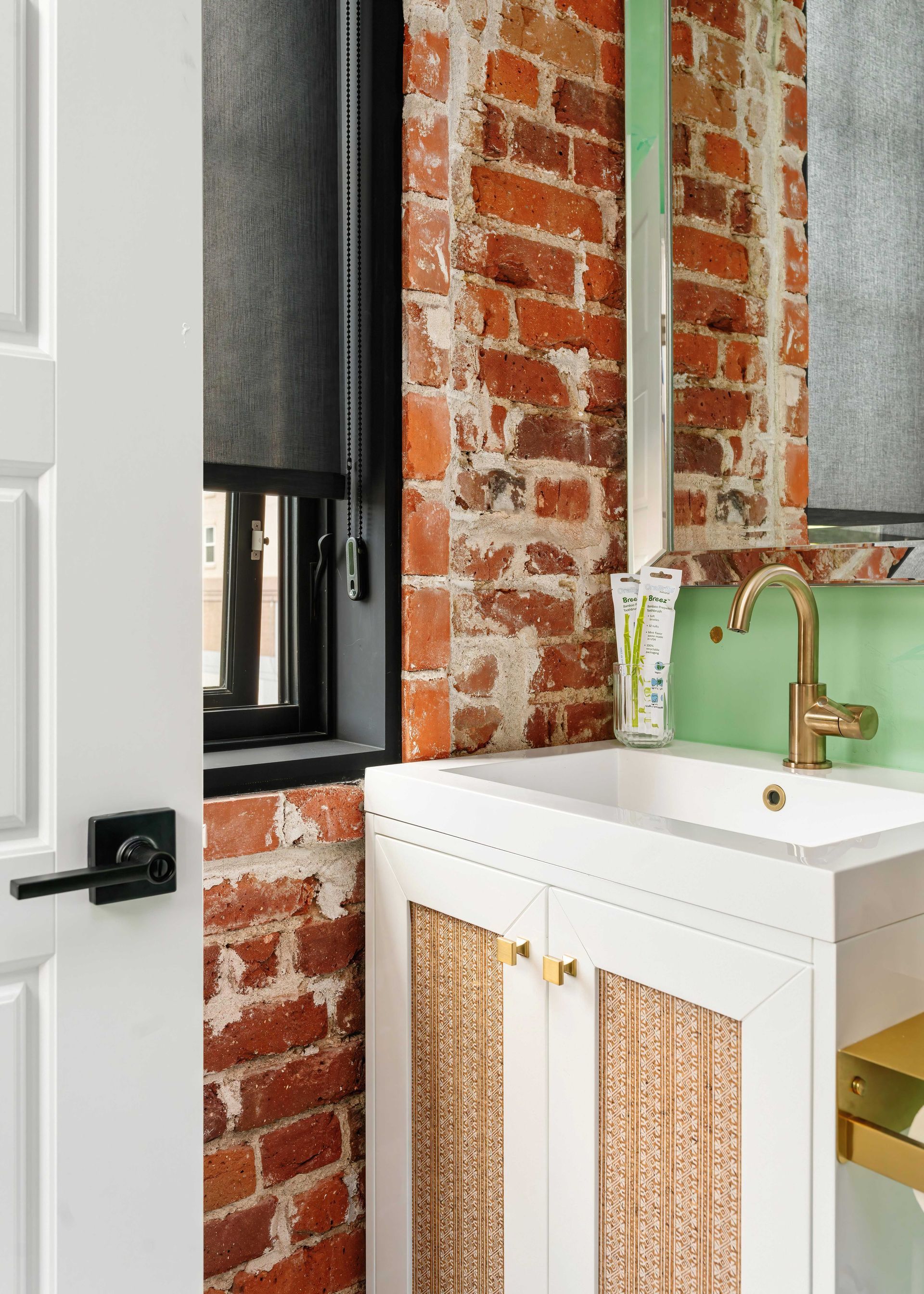 Bathroom vanity with white cabinet, gold faucet, exposed brick wall, and black-framed window