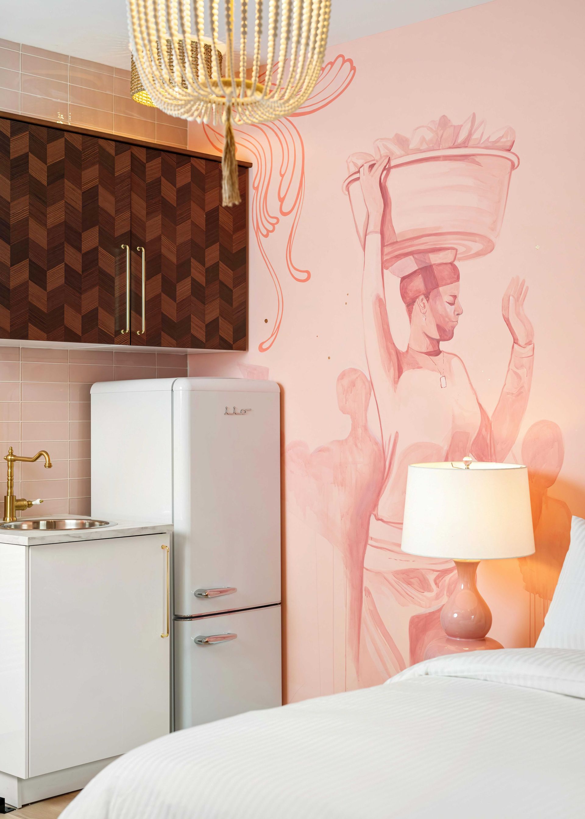Cozy bedroom nook with pink mural, white fridge, lamp, bed, and hanging chandelier