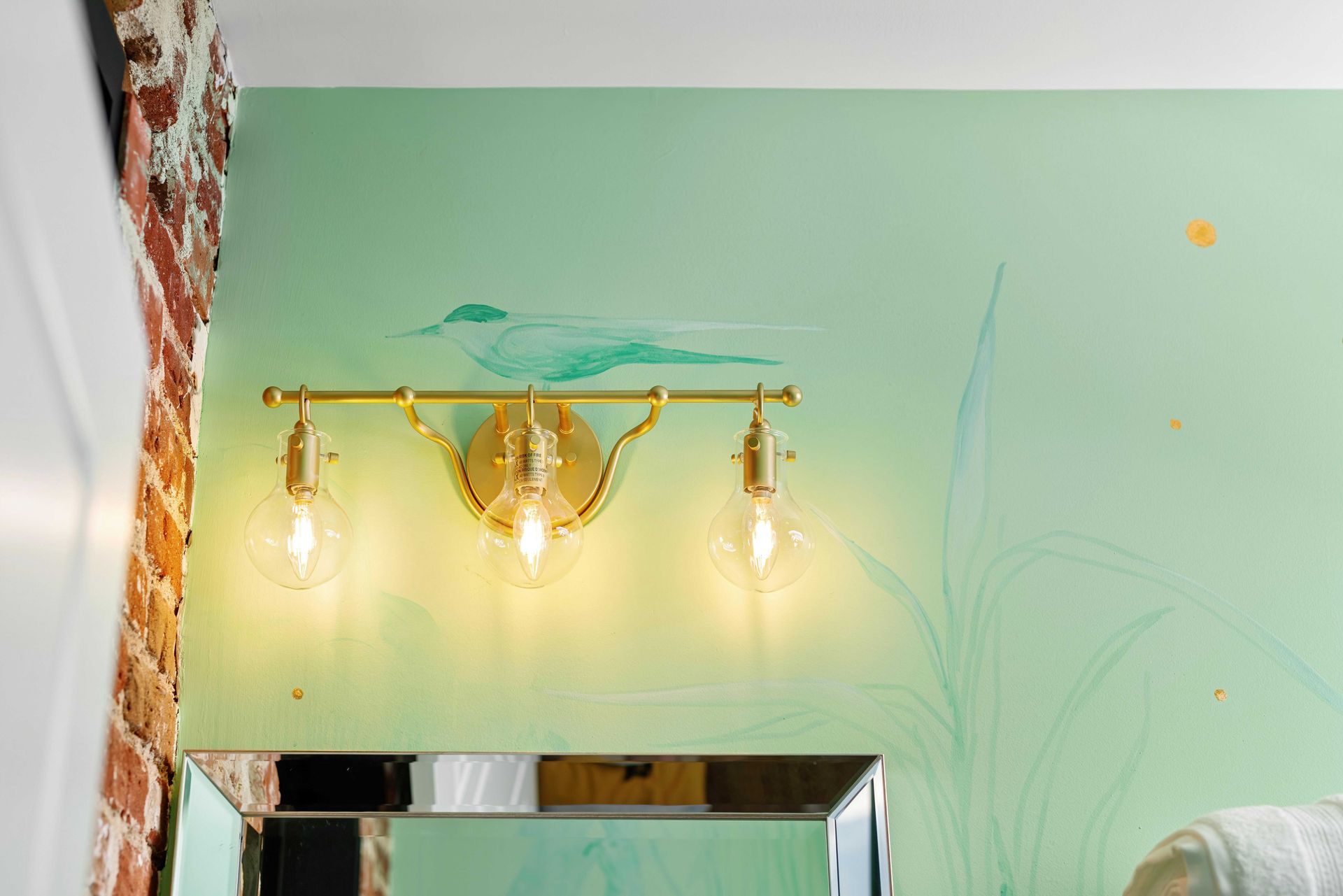 Wall-mounted brass light fixture with three glowing bulbs above a mirror on a mint-green wall.
