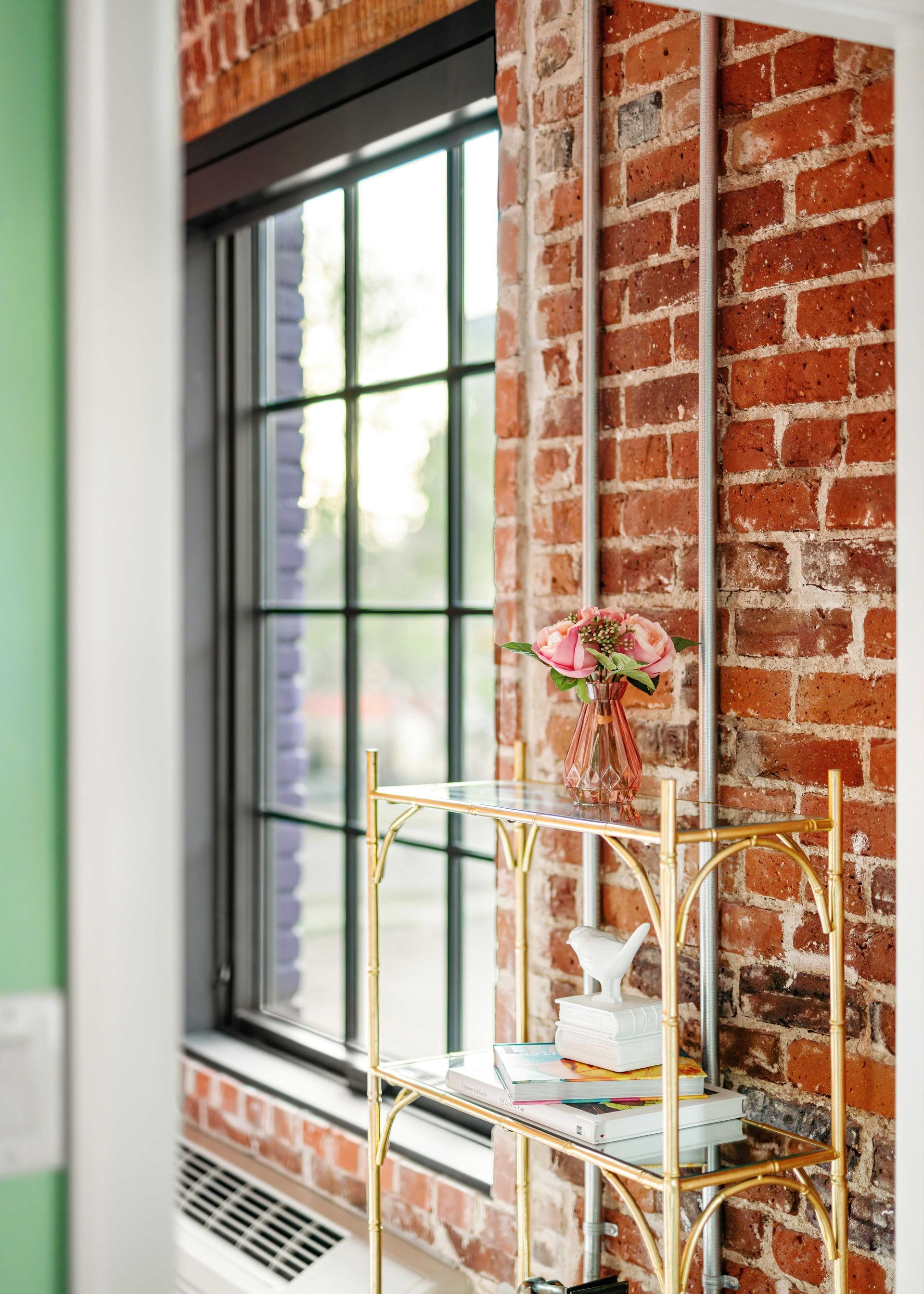 Bright apartment corner with a brick wall, black-framed window, and a gold shelf holding flowers and decor.