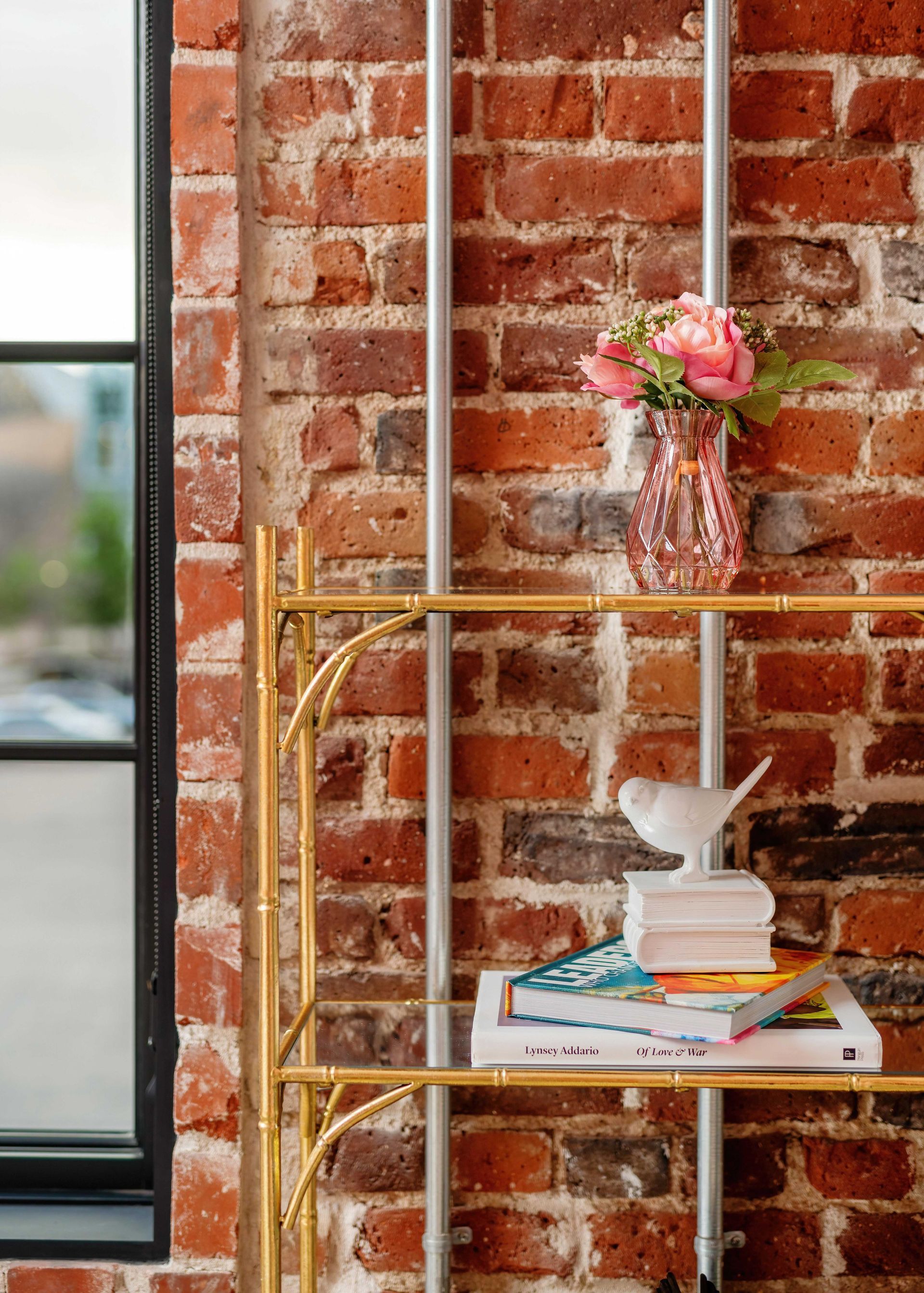 Gold shelf with pink flowers, stacked books, and a white rabbit figurine against a brick wall.