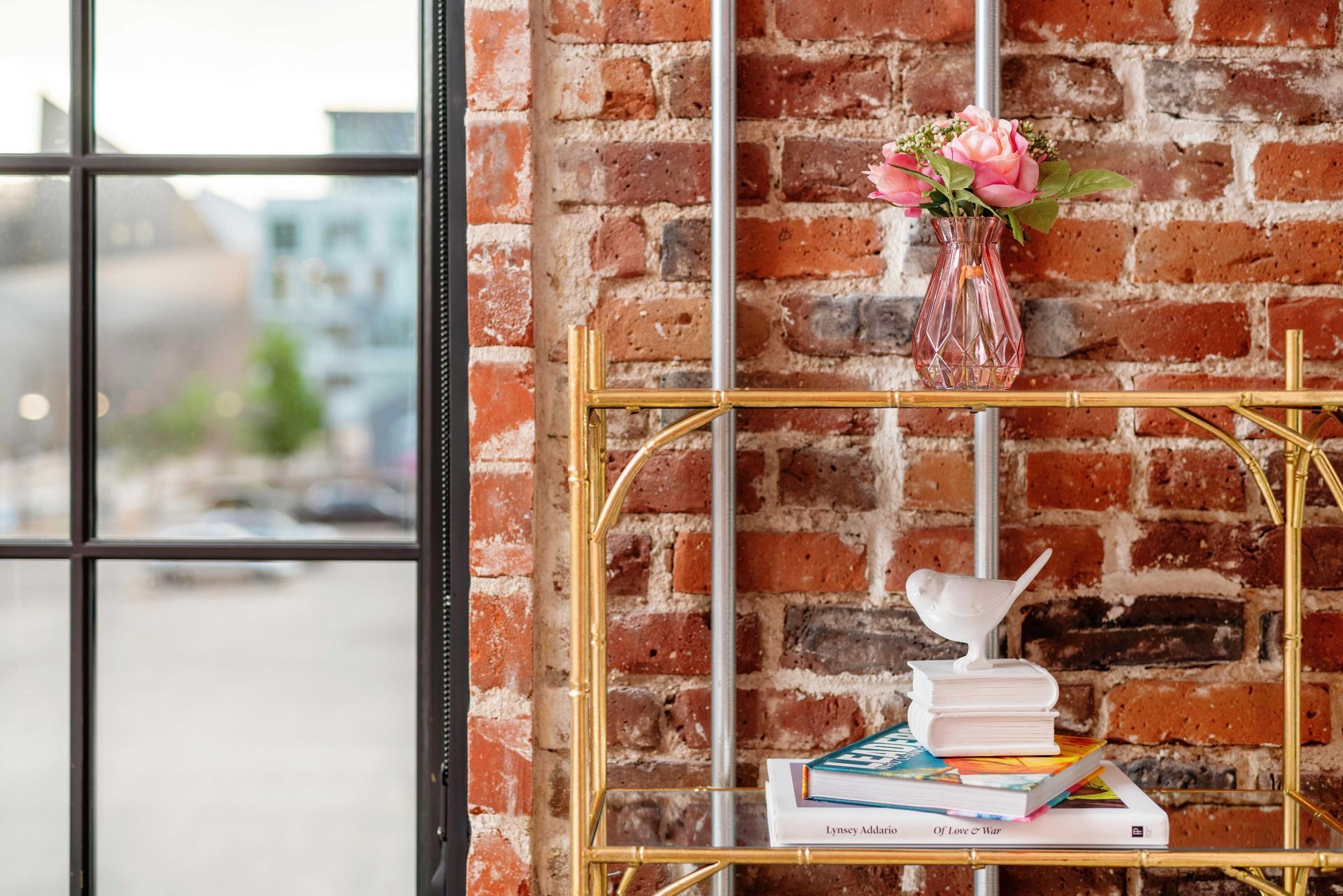 Gold shelf with books and vase of pink flowers beside a black-framed window and red brick wall