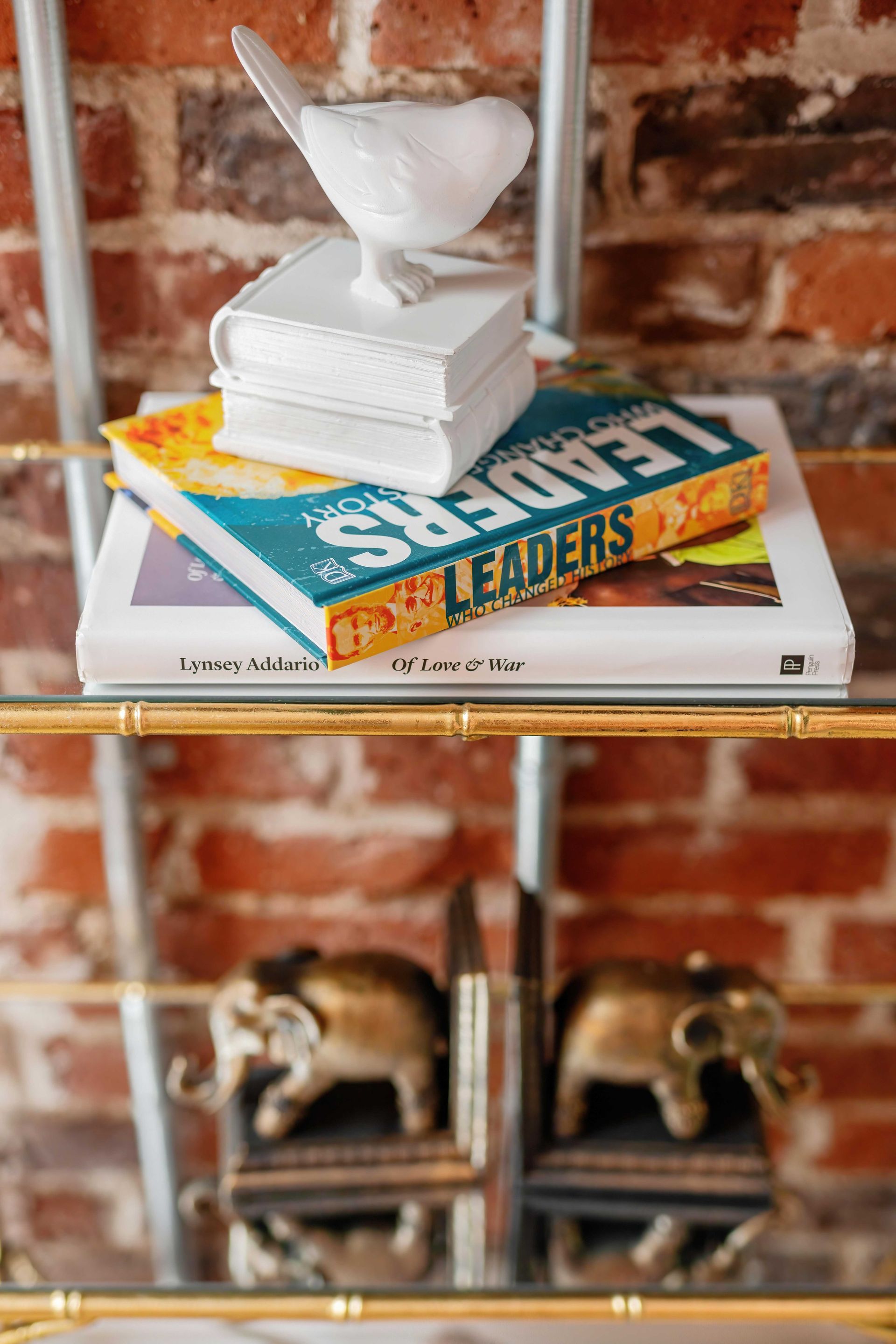 Stacked books with a white decorative object on top, in front of a brick wall and metal shelf decor