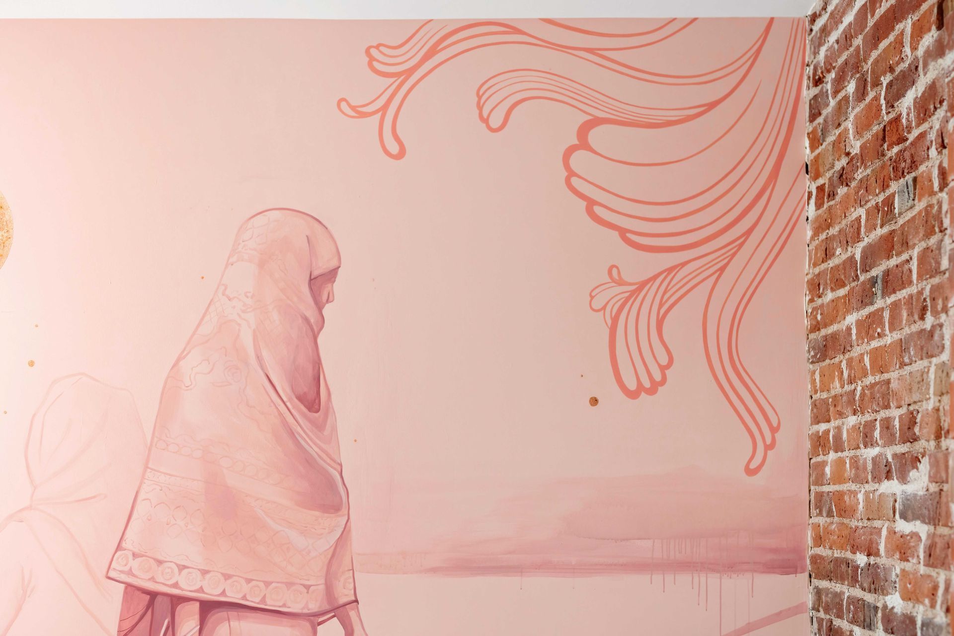 Woman in a pink shawl mural beside a brick wall with flowing coral line art on a pale pink background