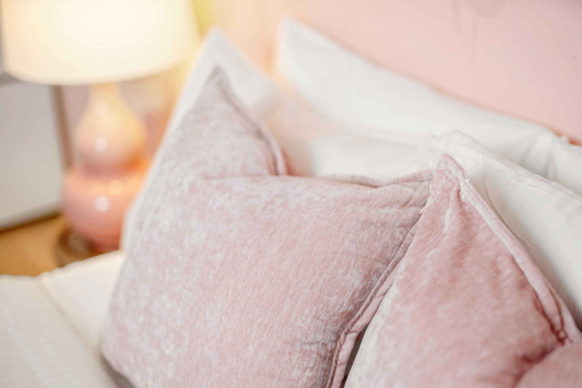 Pink pillows on a bed beside a softly glowing table lamp.