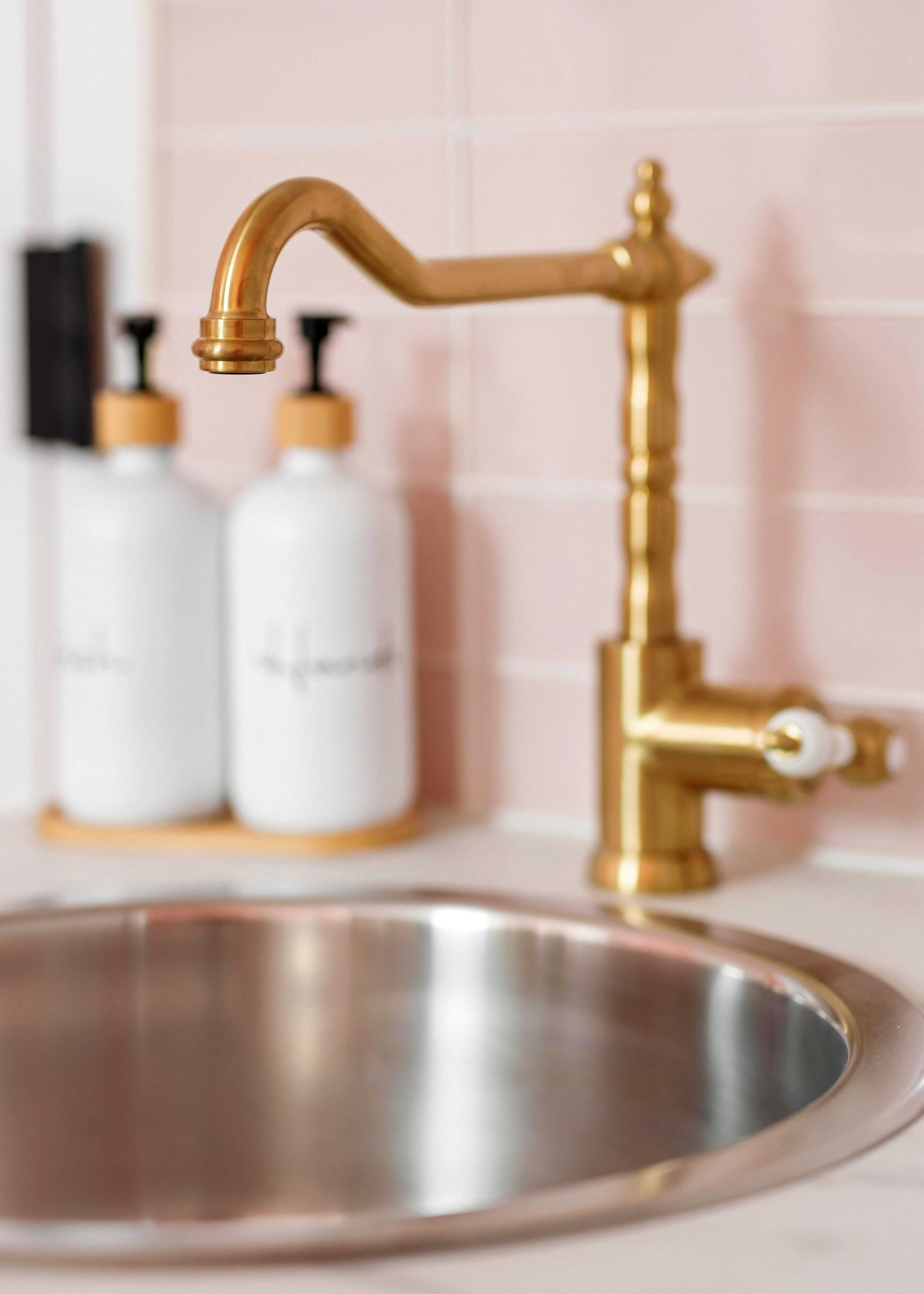 Gold kitchen faucet over a stainless-steel sink with pink tile backsplash and soap bottles in the background
