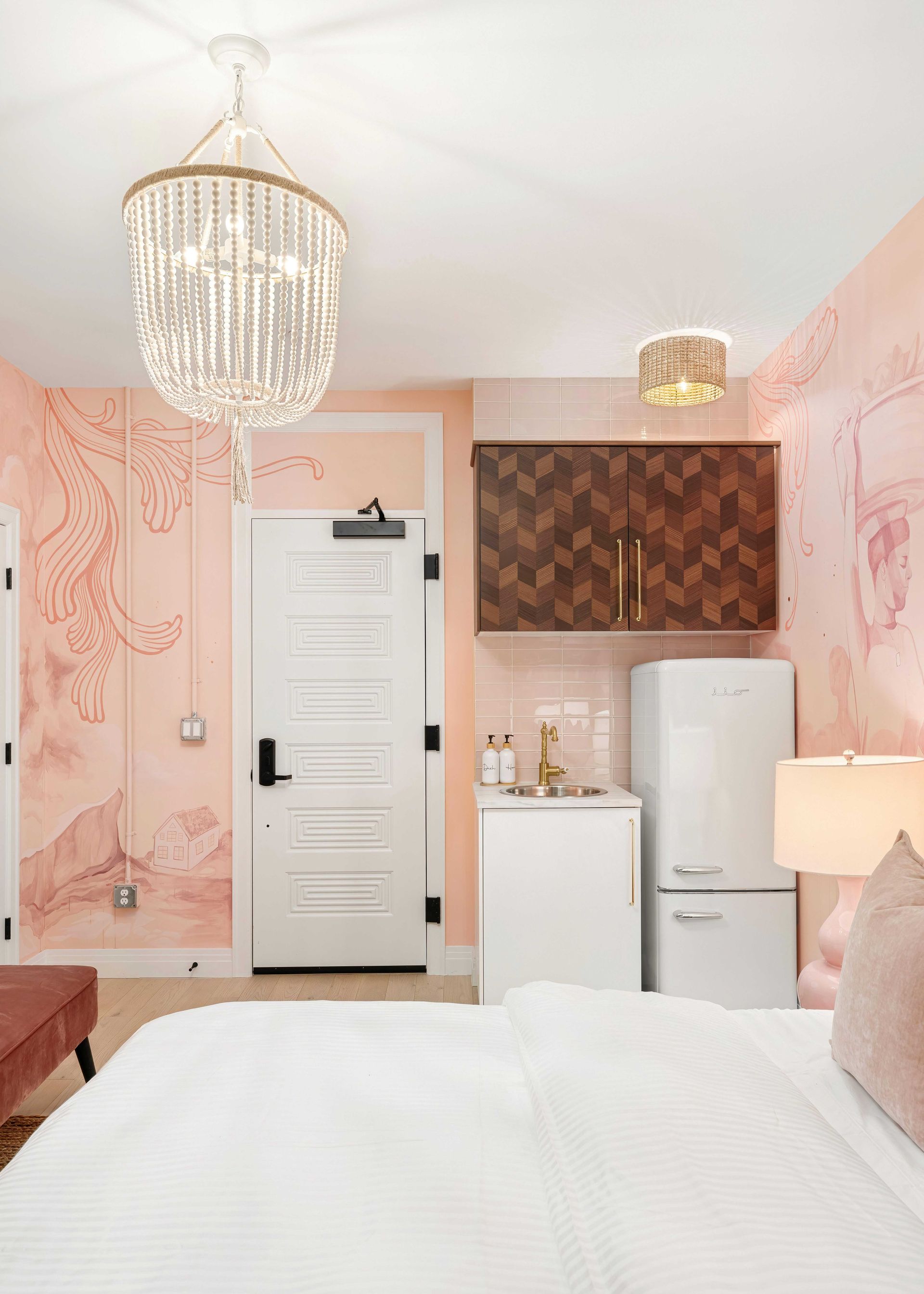 Pink bedroom with crystal chandelier, white bed, and a mini fridge beside the door