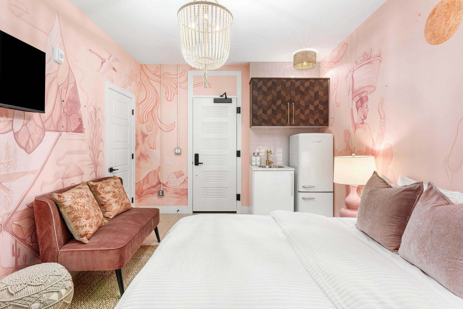 Pink bedroom with white bed, sofa, small fridge, and floral wall art, viewed from the foot of the bed