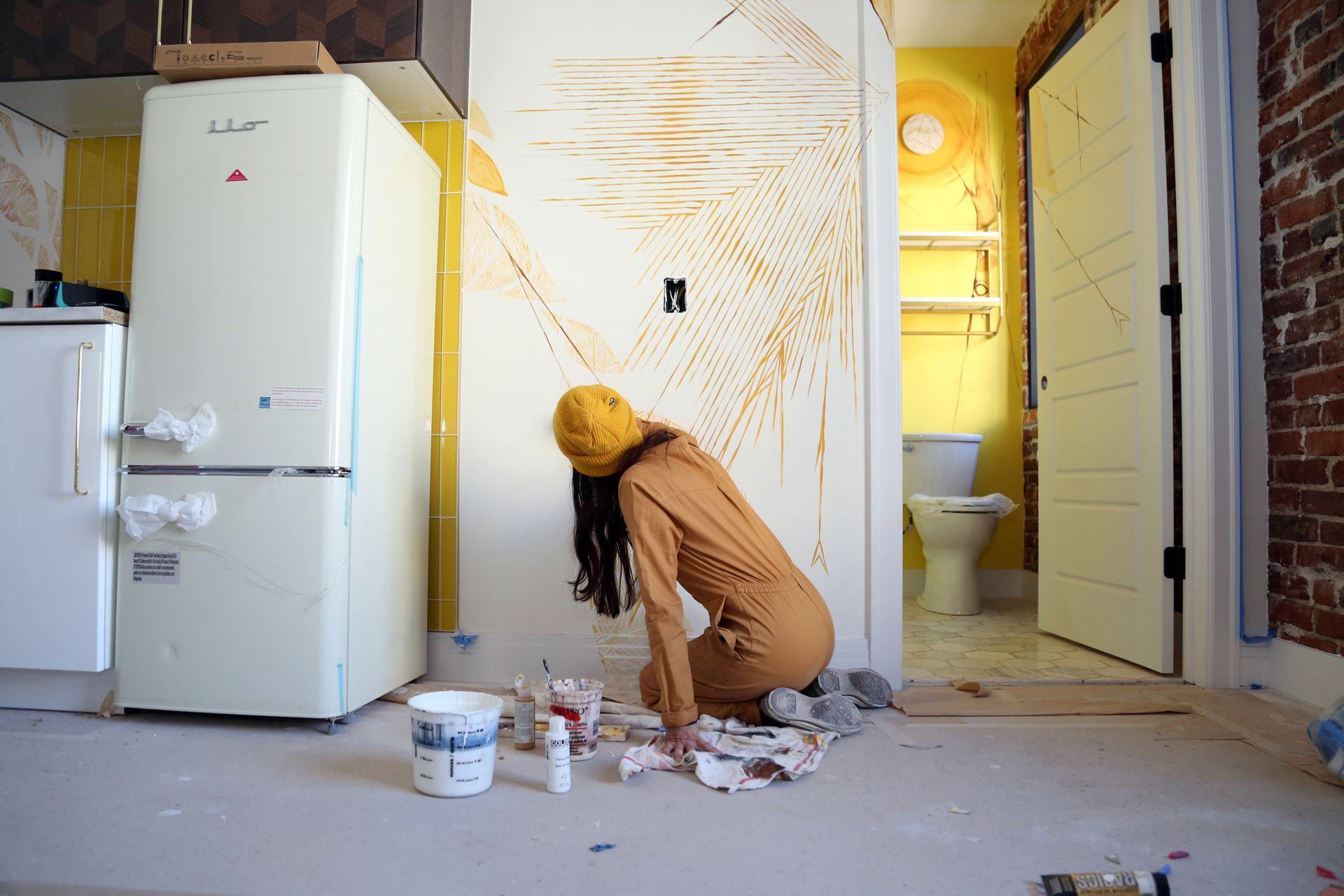 Person painting a wall in a kitchen, with paint cans on the floor and a refrigerator nearby