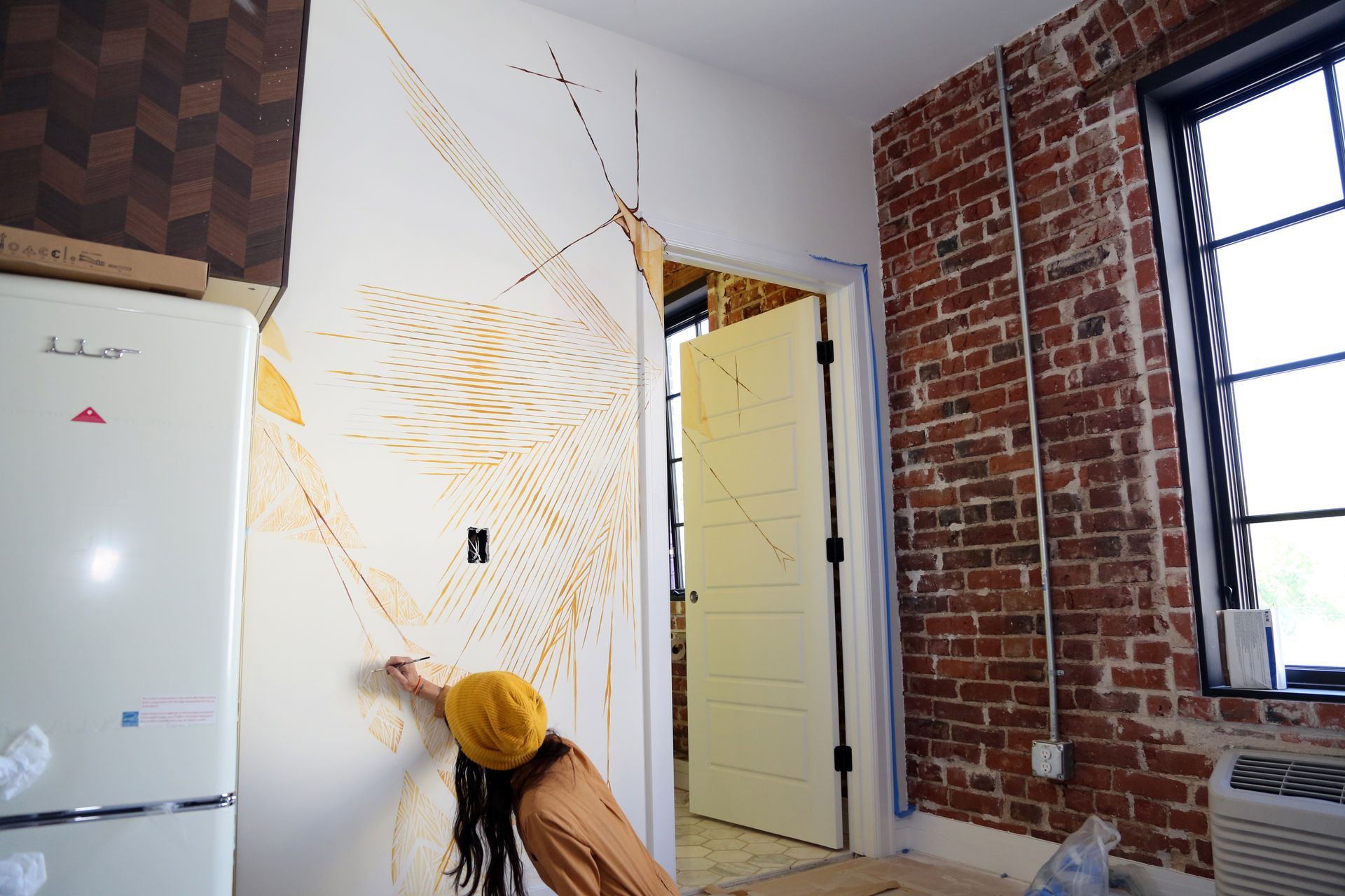 Person painting a white wall with a yellow sunburst mural in a brick-walled room