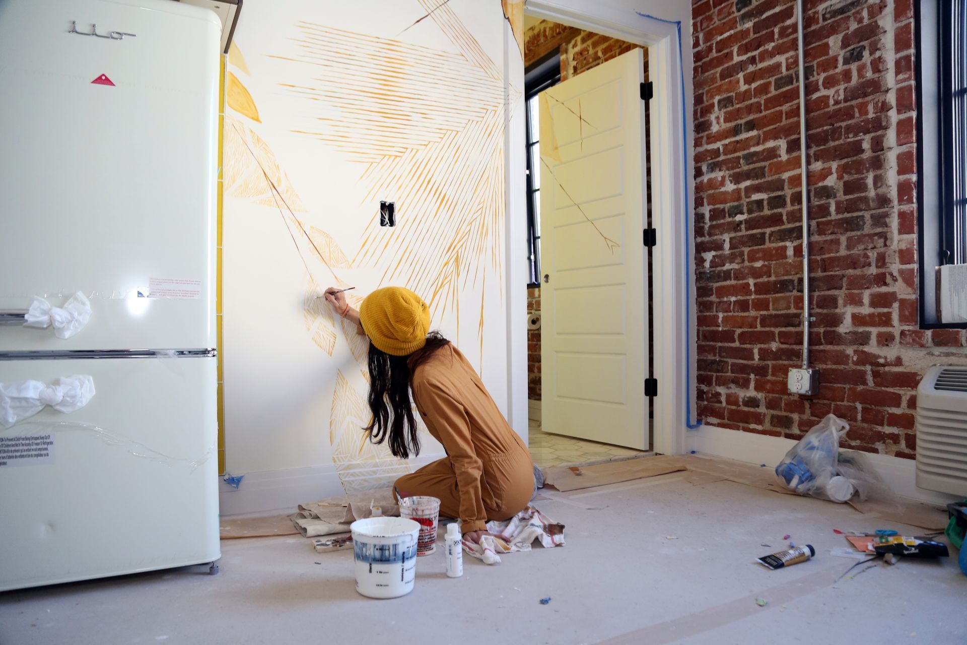 Person painting a wall with a yellow hat in a room under renovation, paint bucket on floor