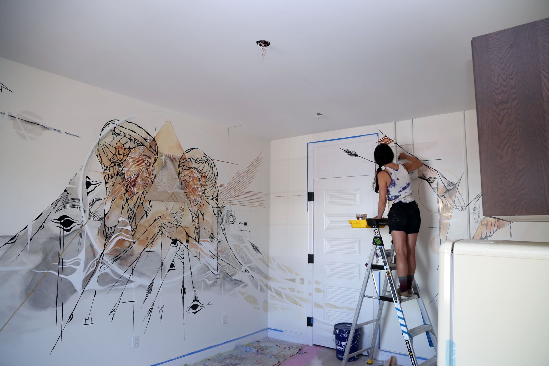 Person painting a large abstract mural on a white wall in a room on a step ladder