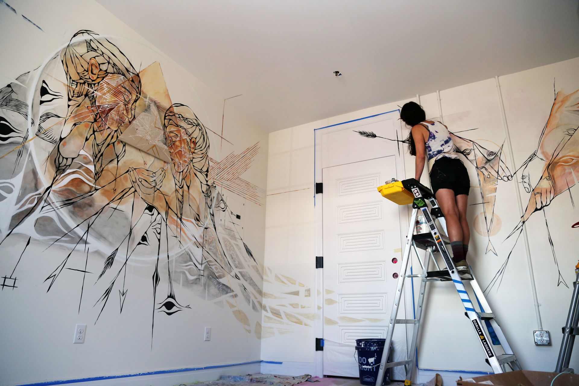 Person on ladder painting a white wall with abstract black and tan mural lines