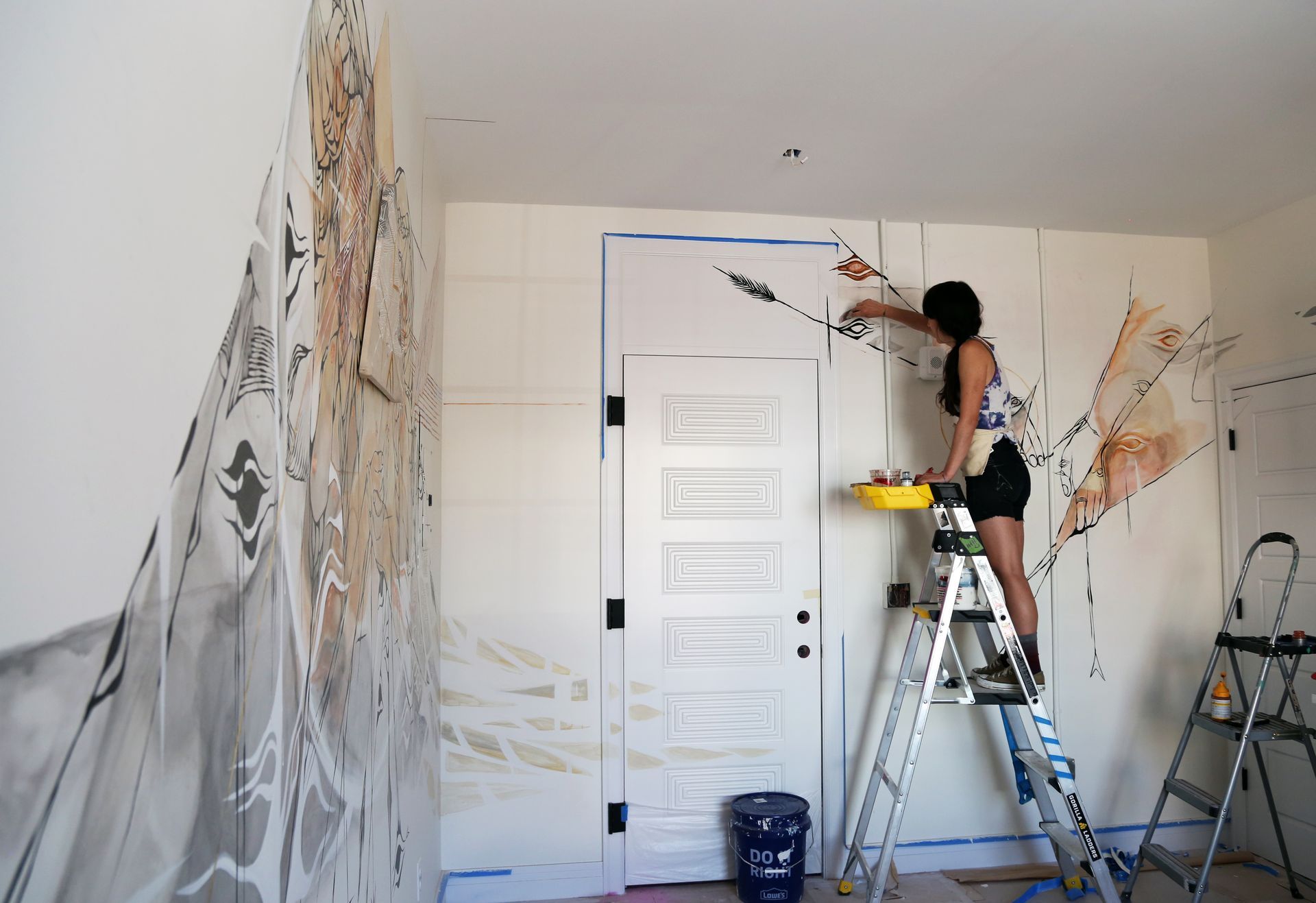 Person painting a room with a ladder, brush, and white walls covered in mural sketches