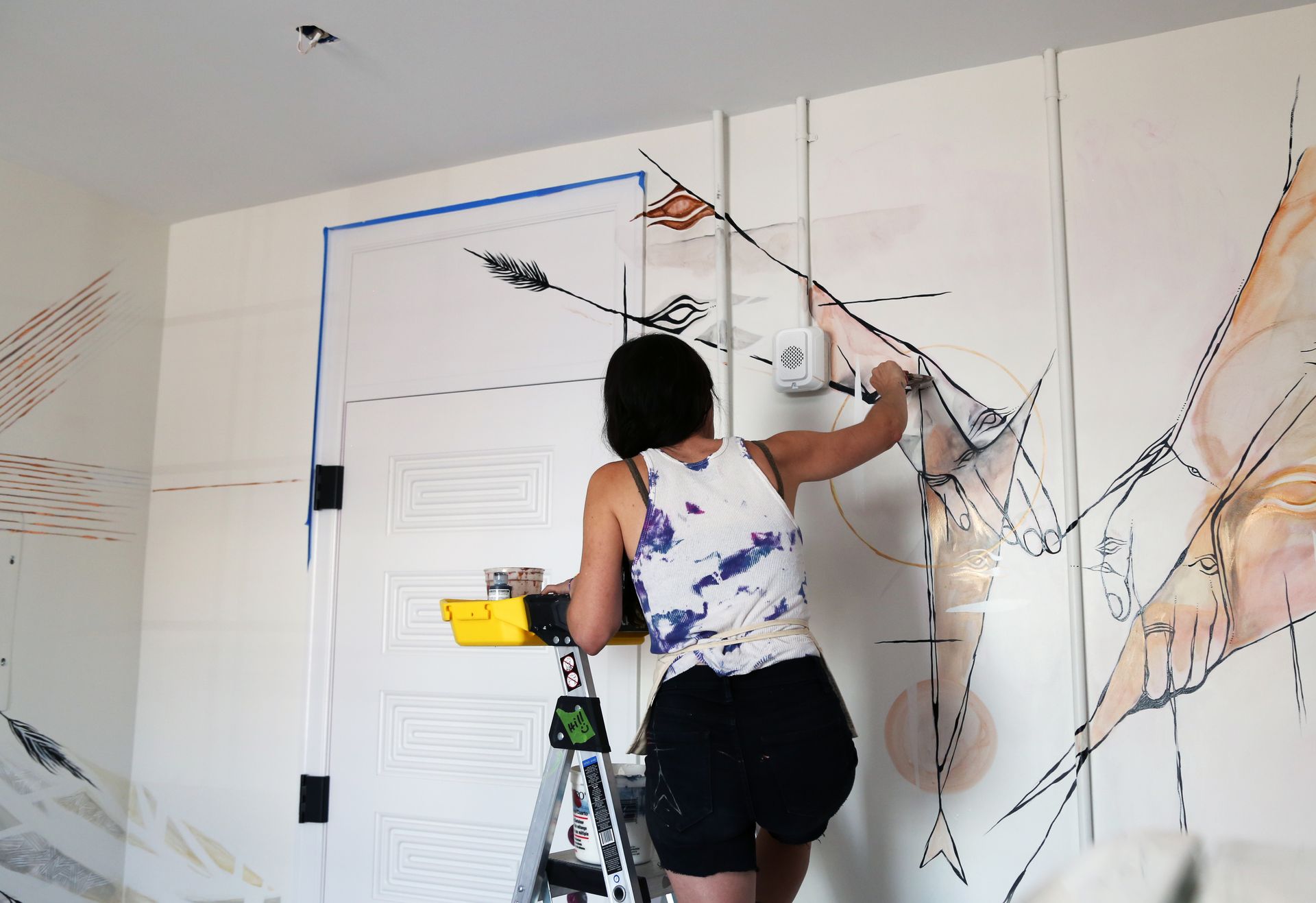 Person painting abstract wall mural on a white wall using a ladder and brush