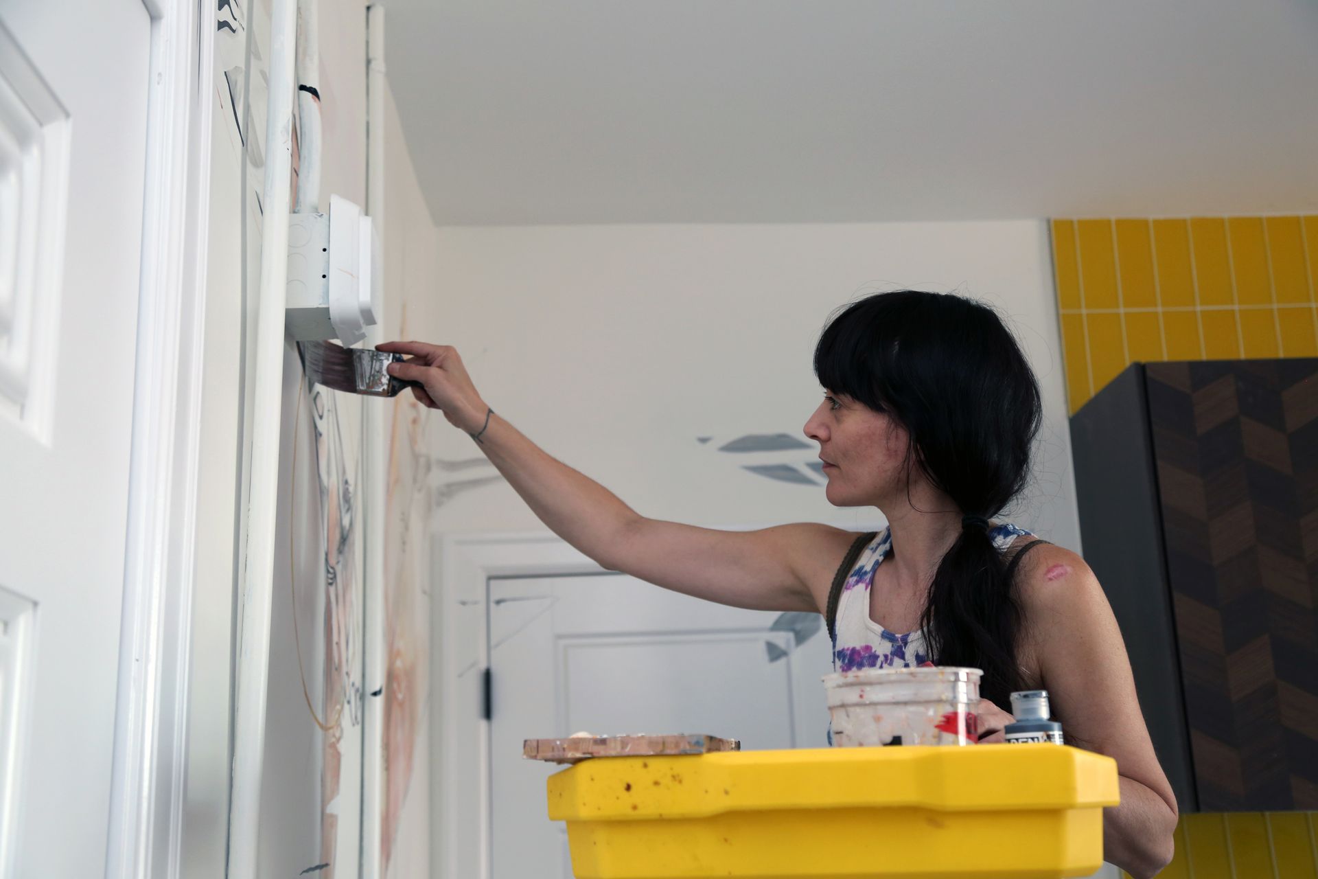 Person painting a white cabinet with a brush, standing beside a yellow bucket in a bright room