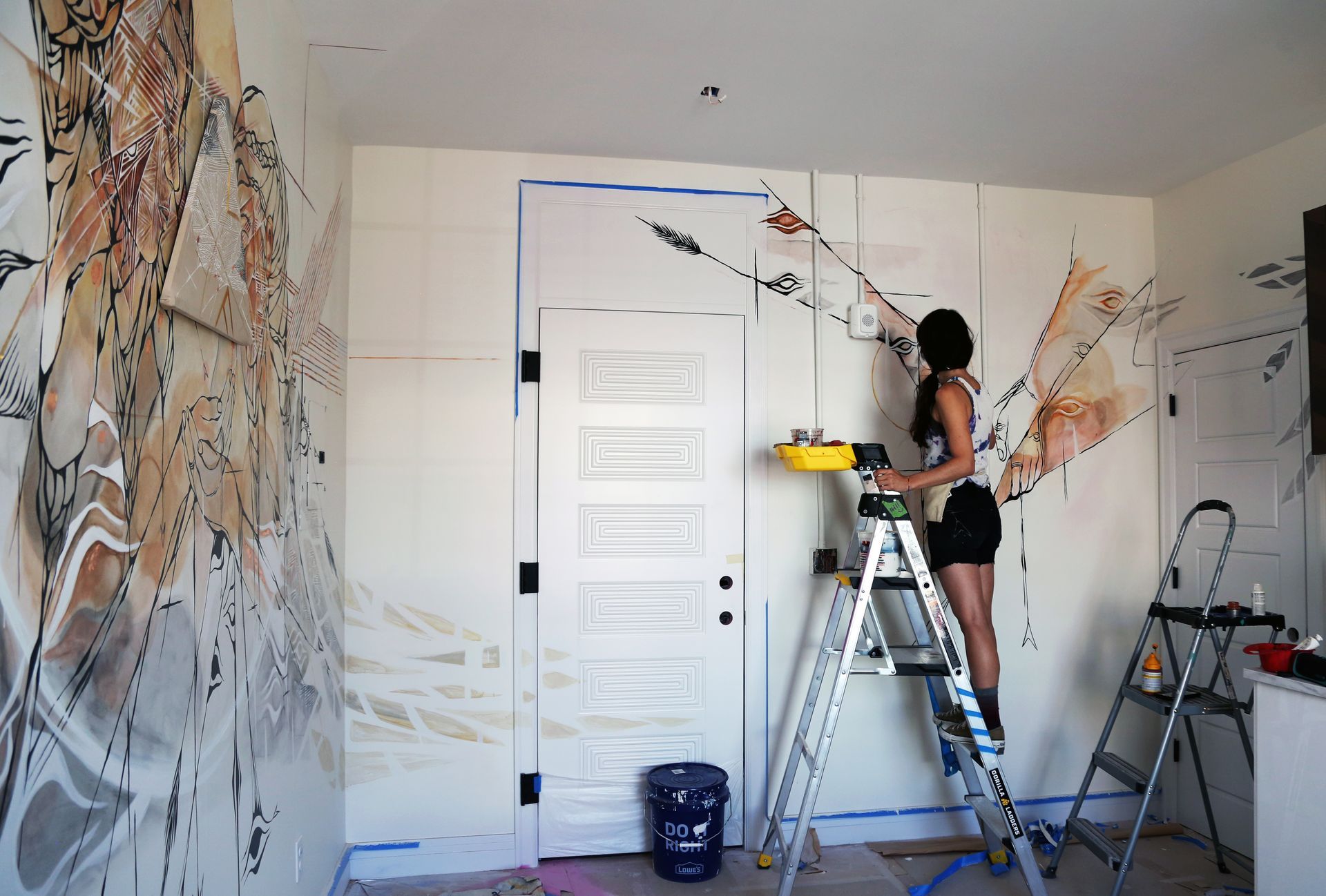 Person painting a white wall beside a door, standing on a step ladder in a room with mural artwork.