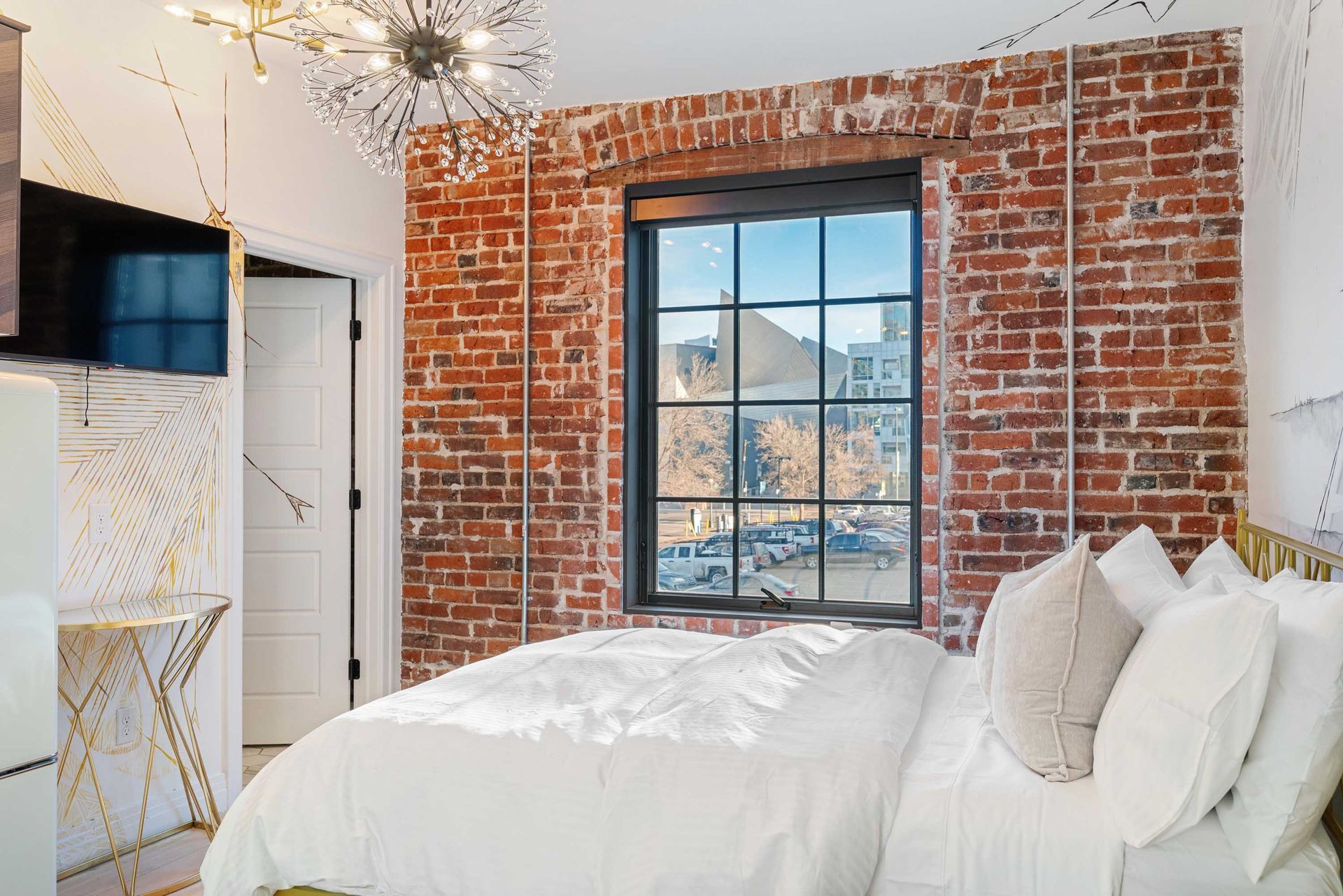 Bright bedroom with exposed brick wall, large window, white bedding, and wall-mounted TV