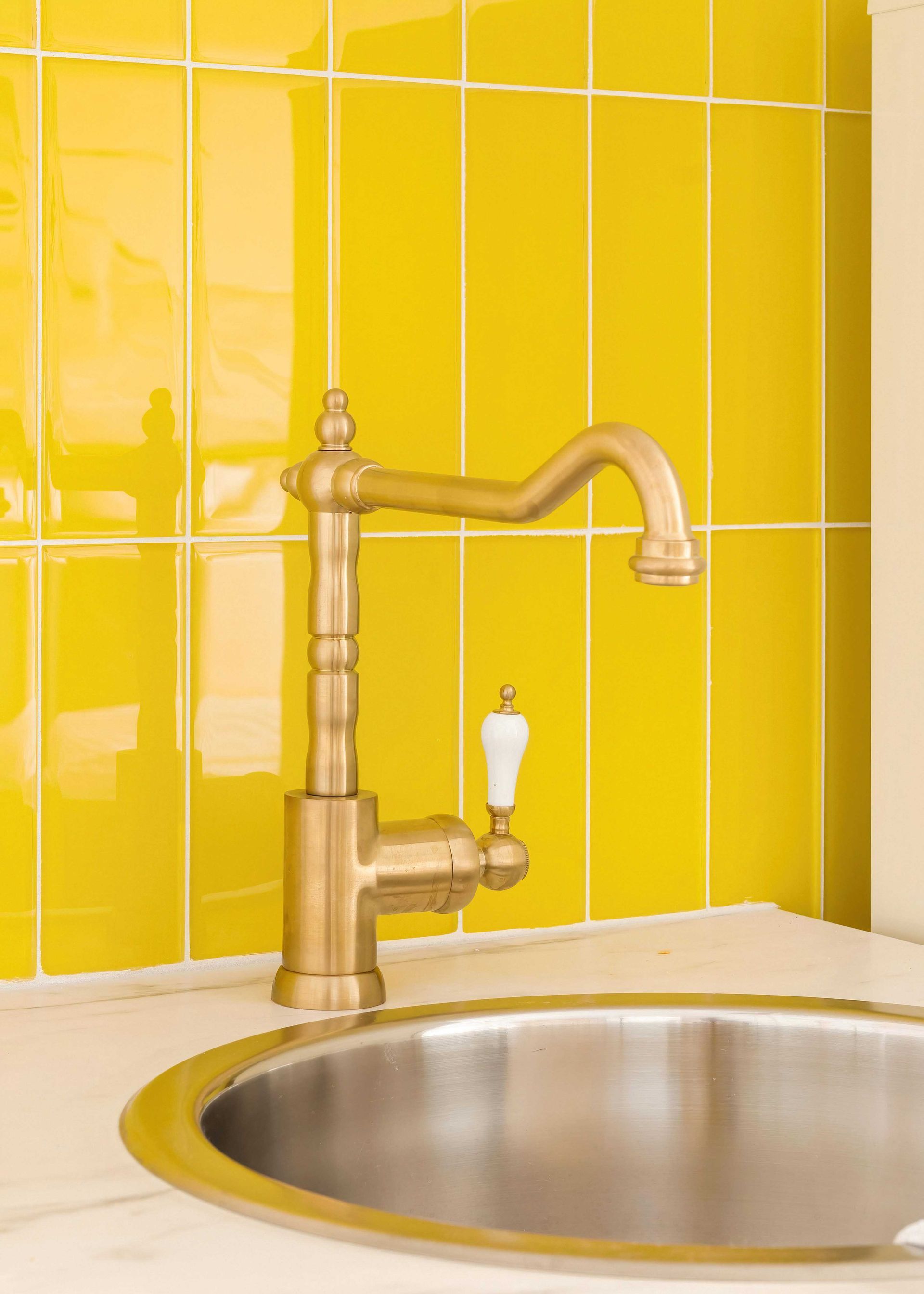 Yellow tiled kitchen wall with a brass faucet over a stainless steel sink