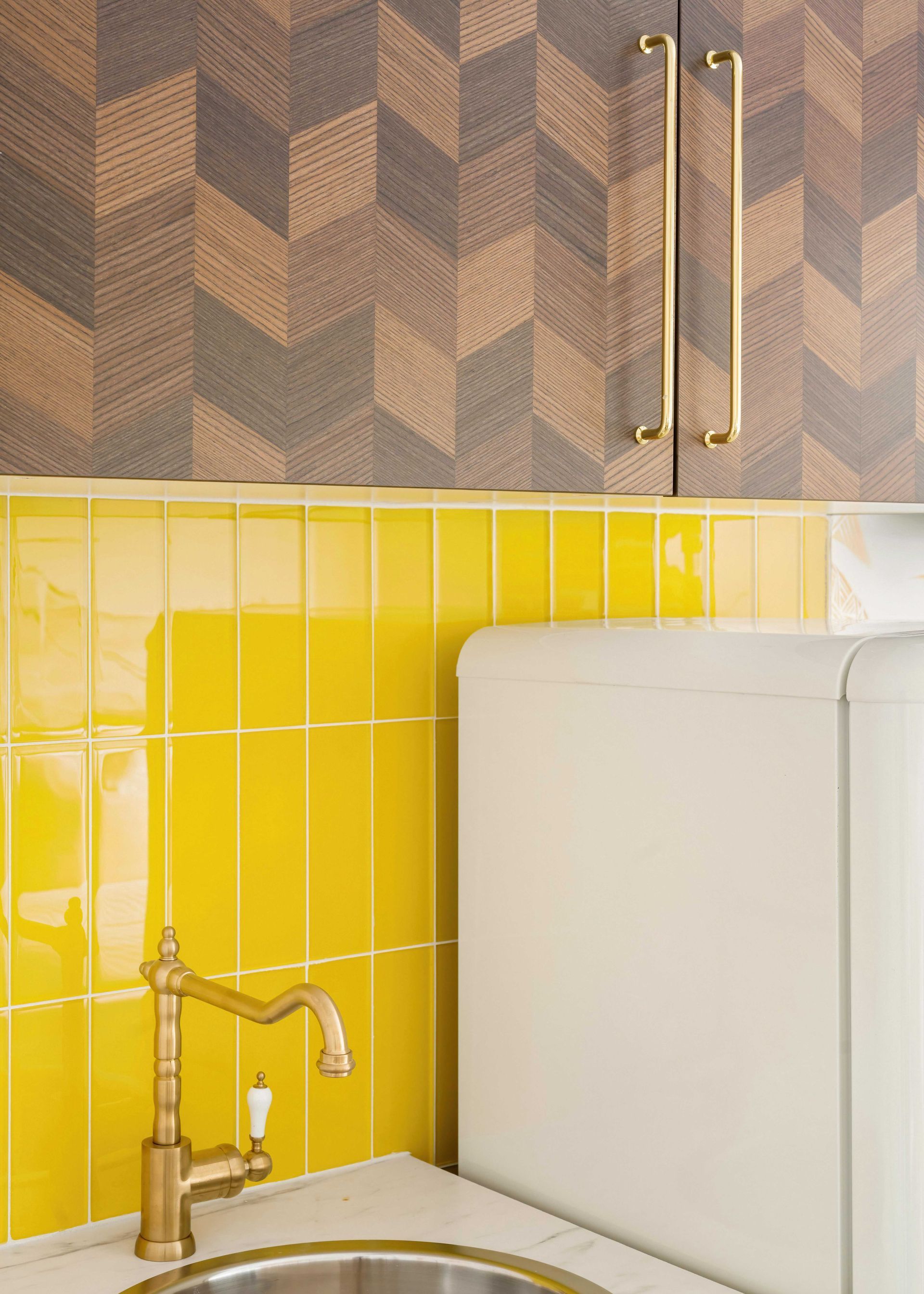 Yellow-tiled kitchen corner with brass faucet, white appliance, and geometric wallpaper.