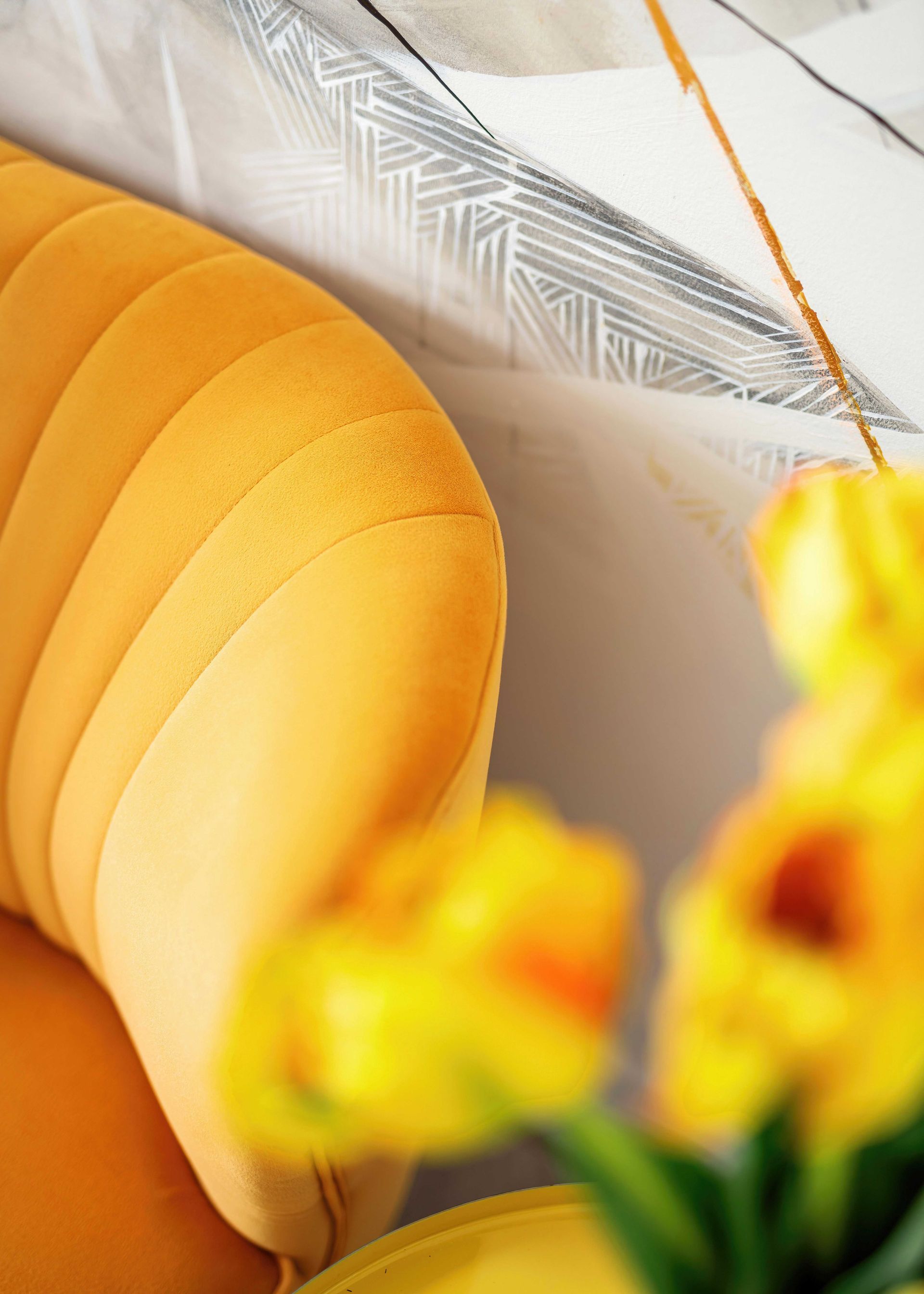 Yellow velvet chair beside a white patterned cushion, with blurred yellow flowers in the foreground