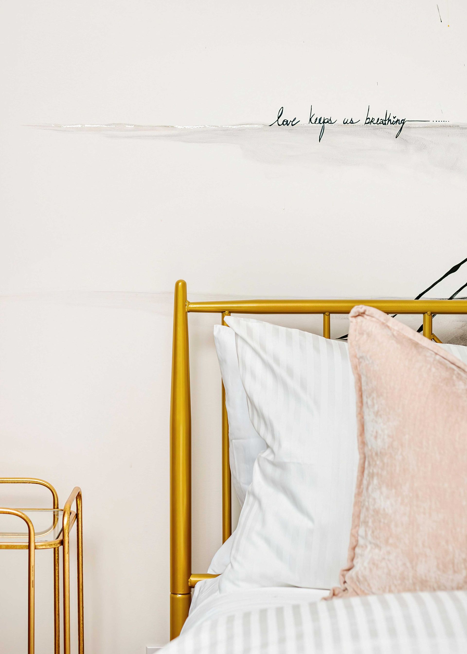 Minimal bedroom corner with a yellow metal bed frame, white and blush pillows, and a small gold side table.
