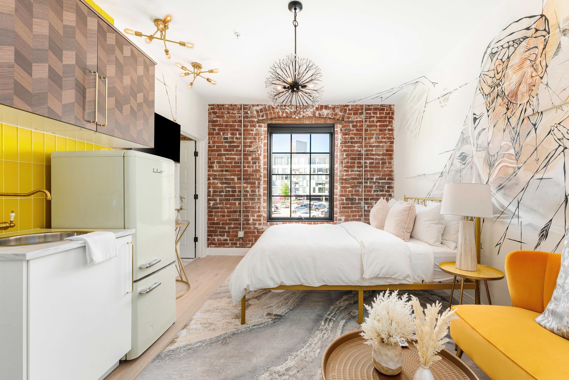 Bright loft bedroom with exposed brick wall, white bedding, yellow kitchen accents, and a mustard chair