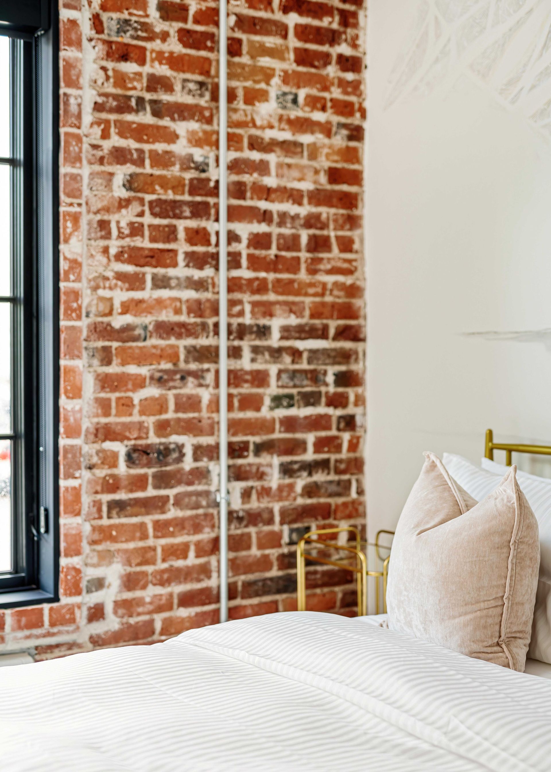 Bedroom with exposed brick wall, black window frame, and white bed with cream pillows