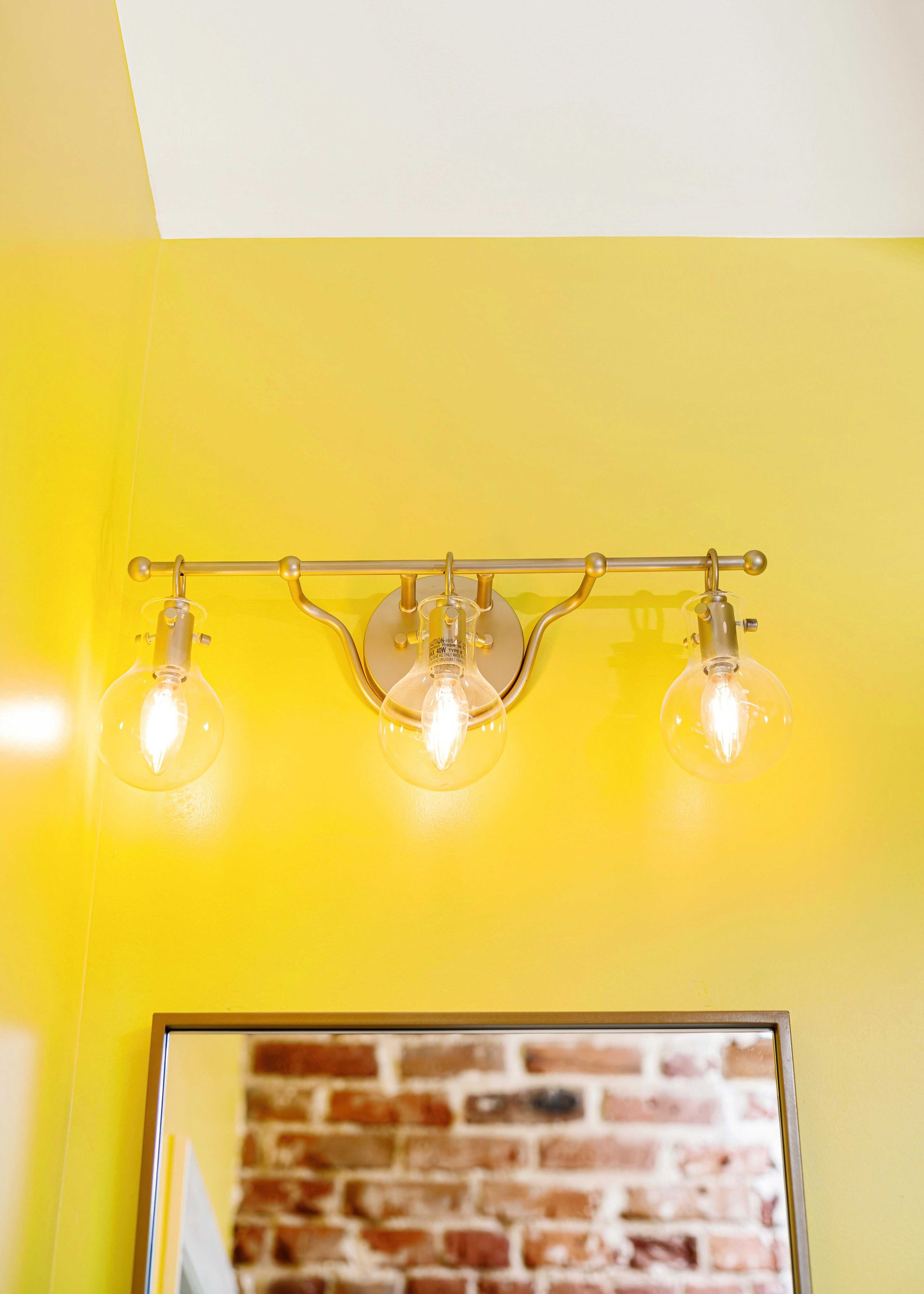 Wall-mounted brass light fixture with three glowing bulbs above a brick-framed mirror on a yellow wall
