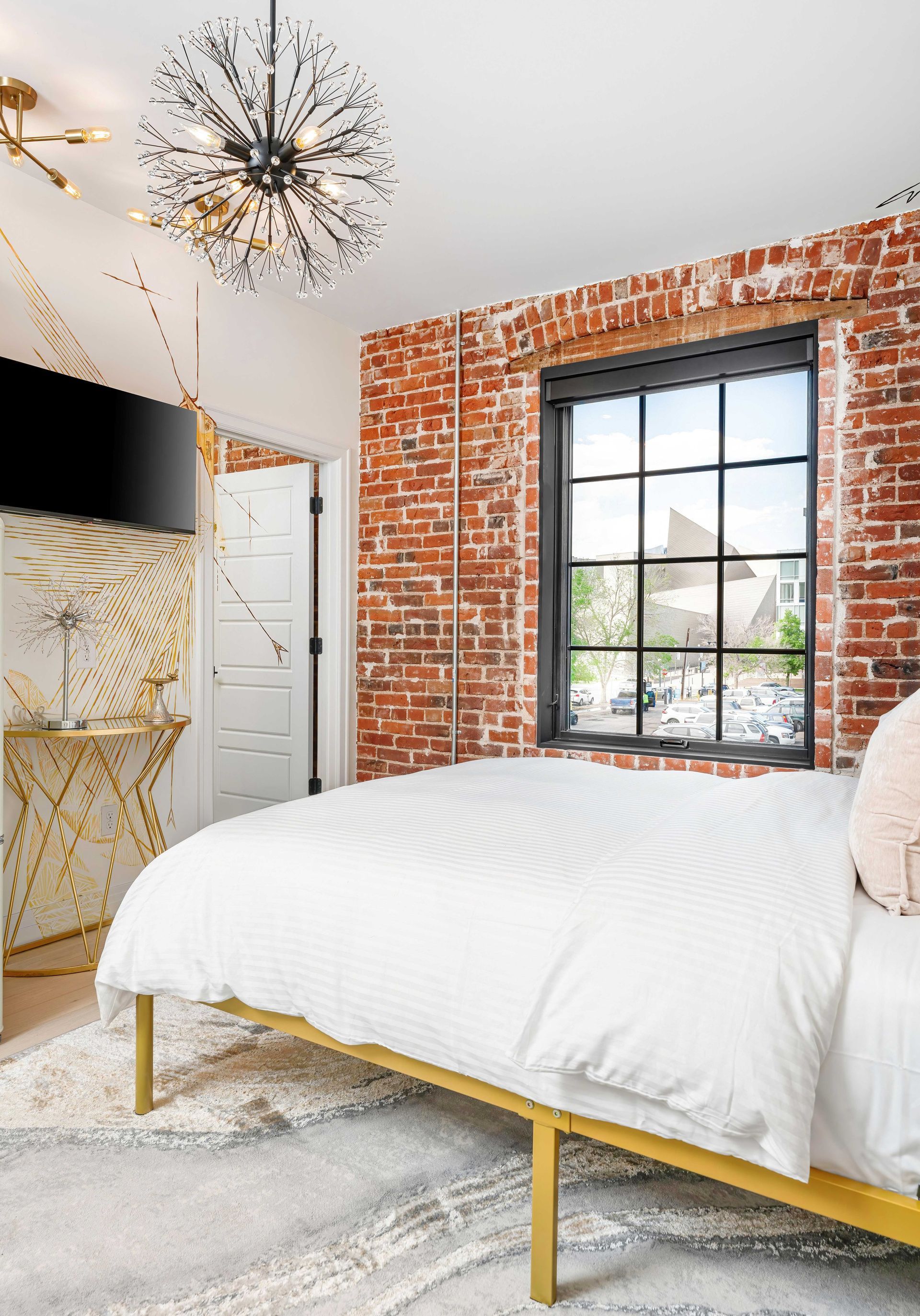 Bright bedroom with exposed brick wall, large black-framed window, white bed, and modern chandelier.
