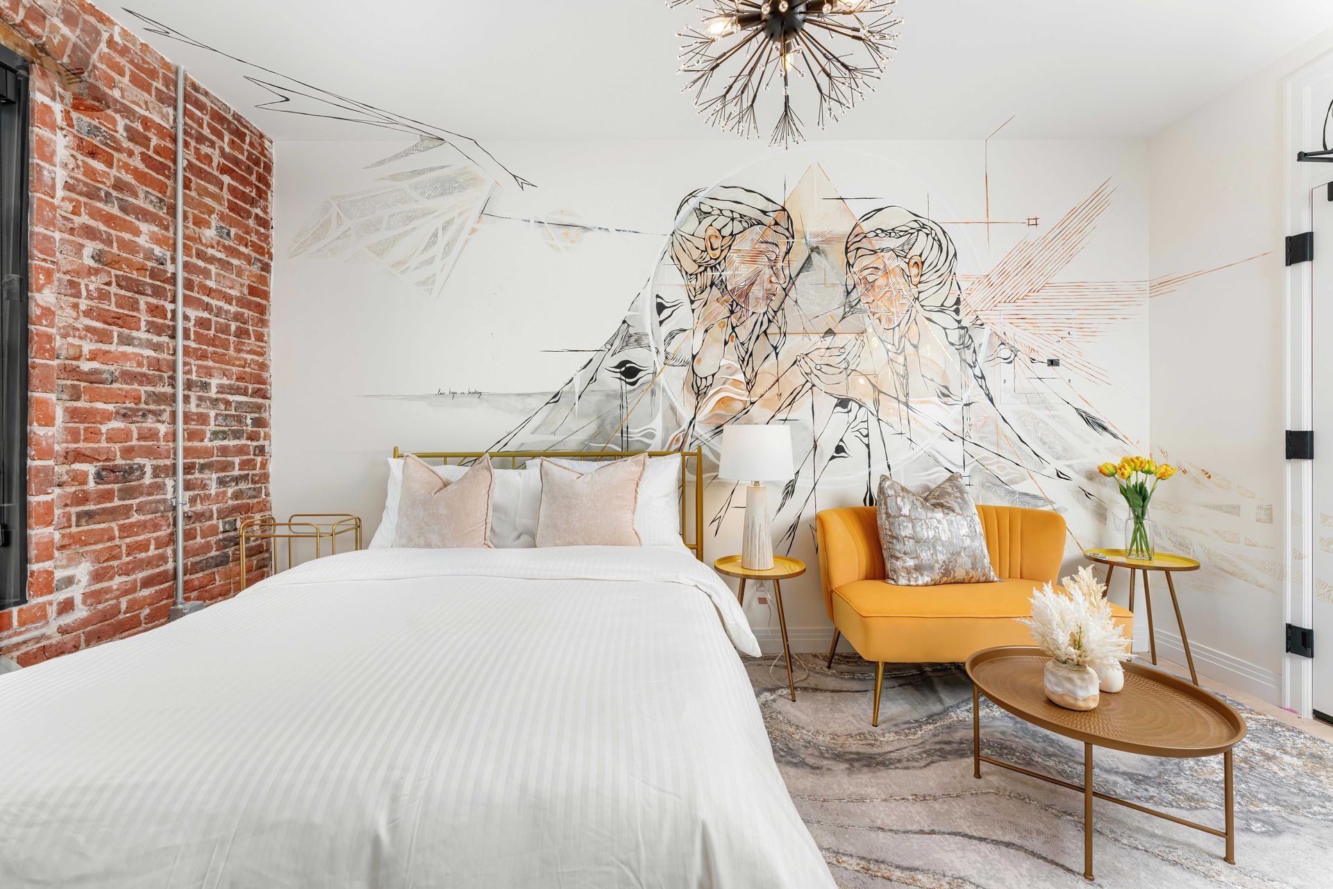Bright modern bedroom with exposed brick wall, white bedding, mustard chair, and floral wall mural