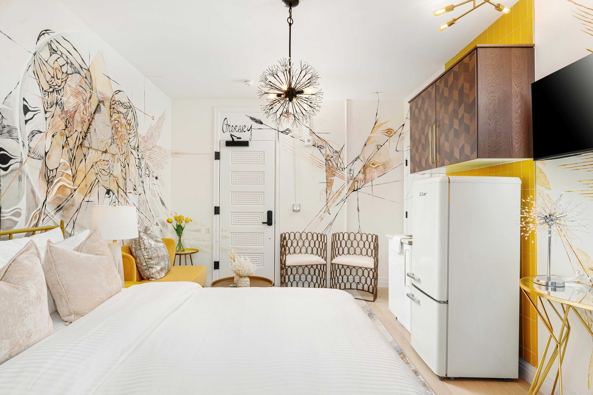 Bright white bedroom with gold abstract wall art, a bed, chair, dresser, and small refrigerator