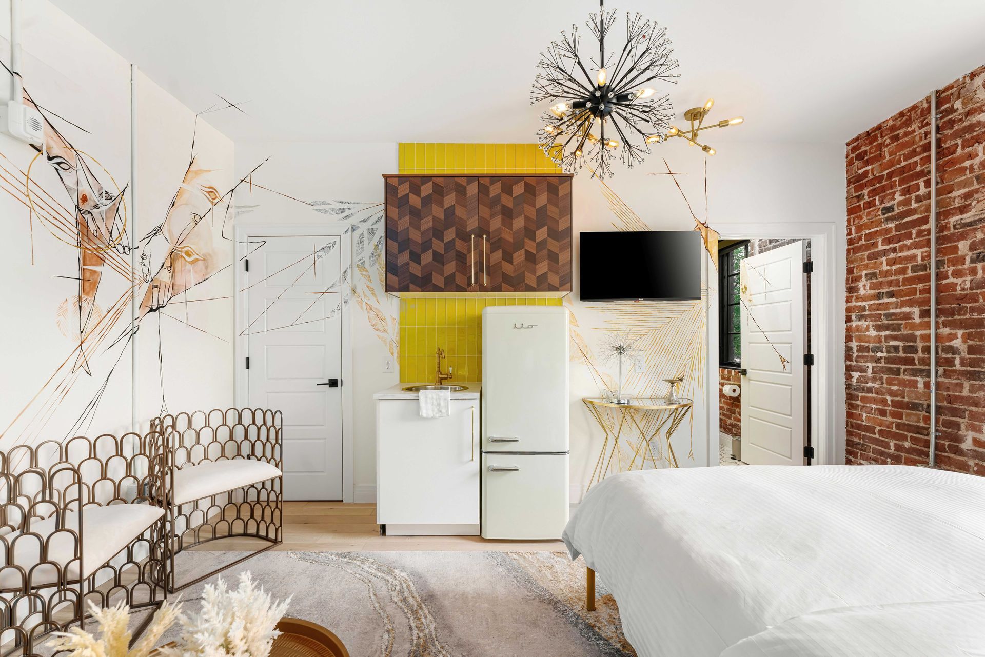 Bright eclectic bedroom with white bed, patterned chair, yellow cabinet, brick accent wall, and modern chandelier