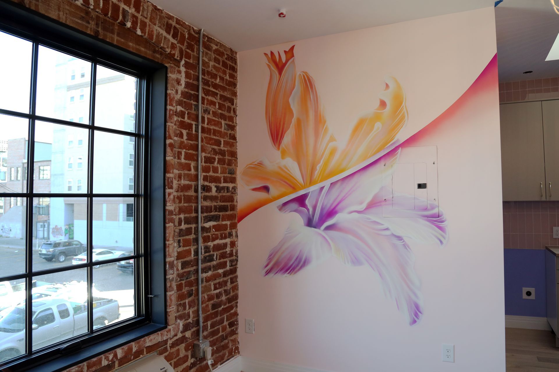 Abstract orange, purple, and pink mural on a white wall beside a brick window in a bright room