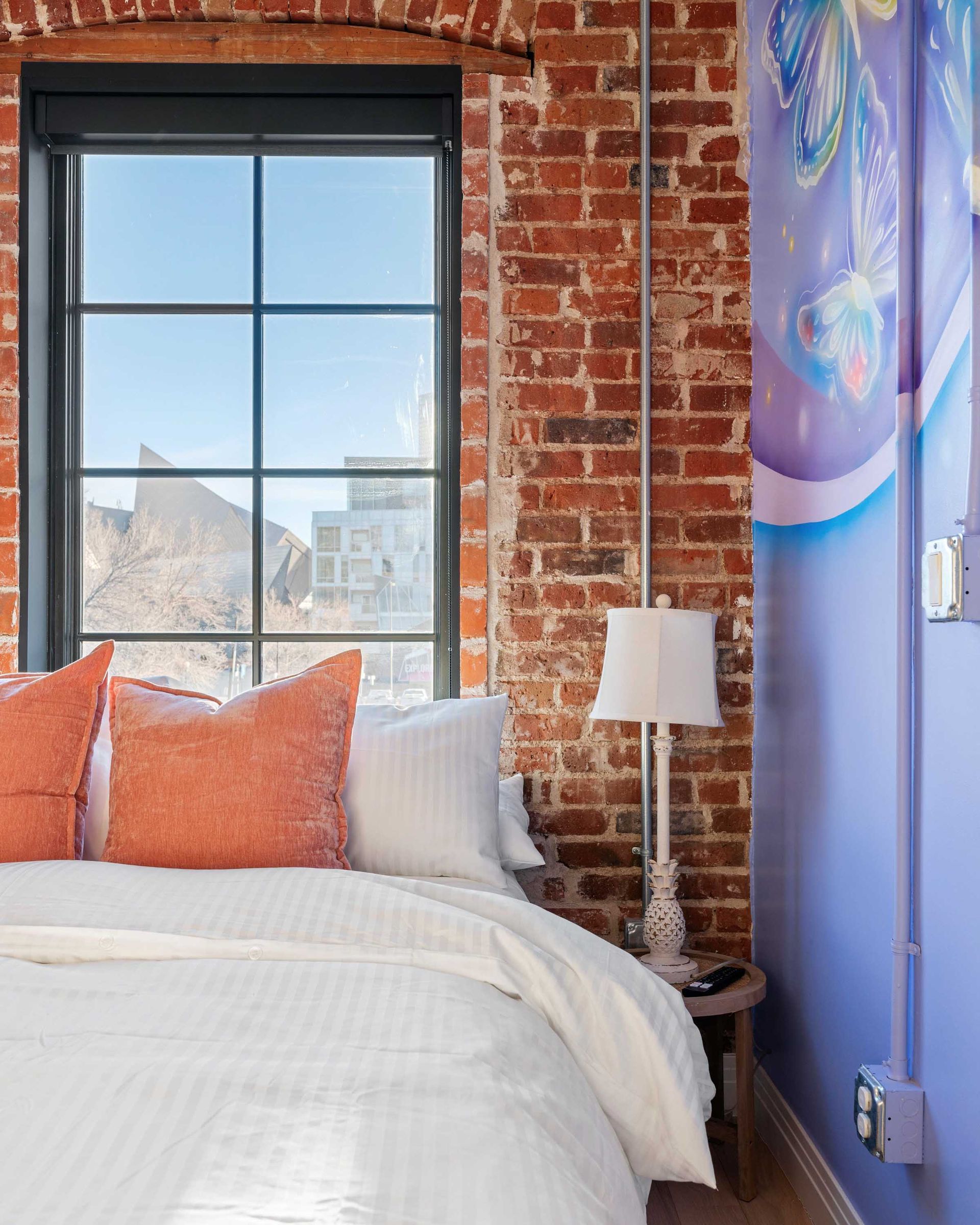 Cozy bedroom with brick wall, large window, white bed, orange pillows, and a small lamp