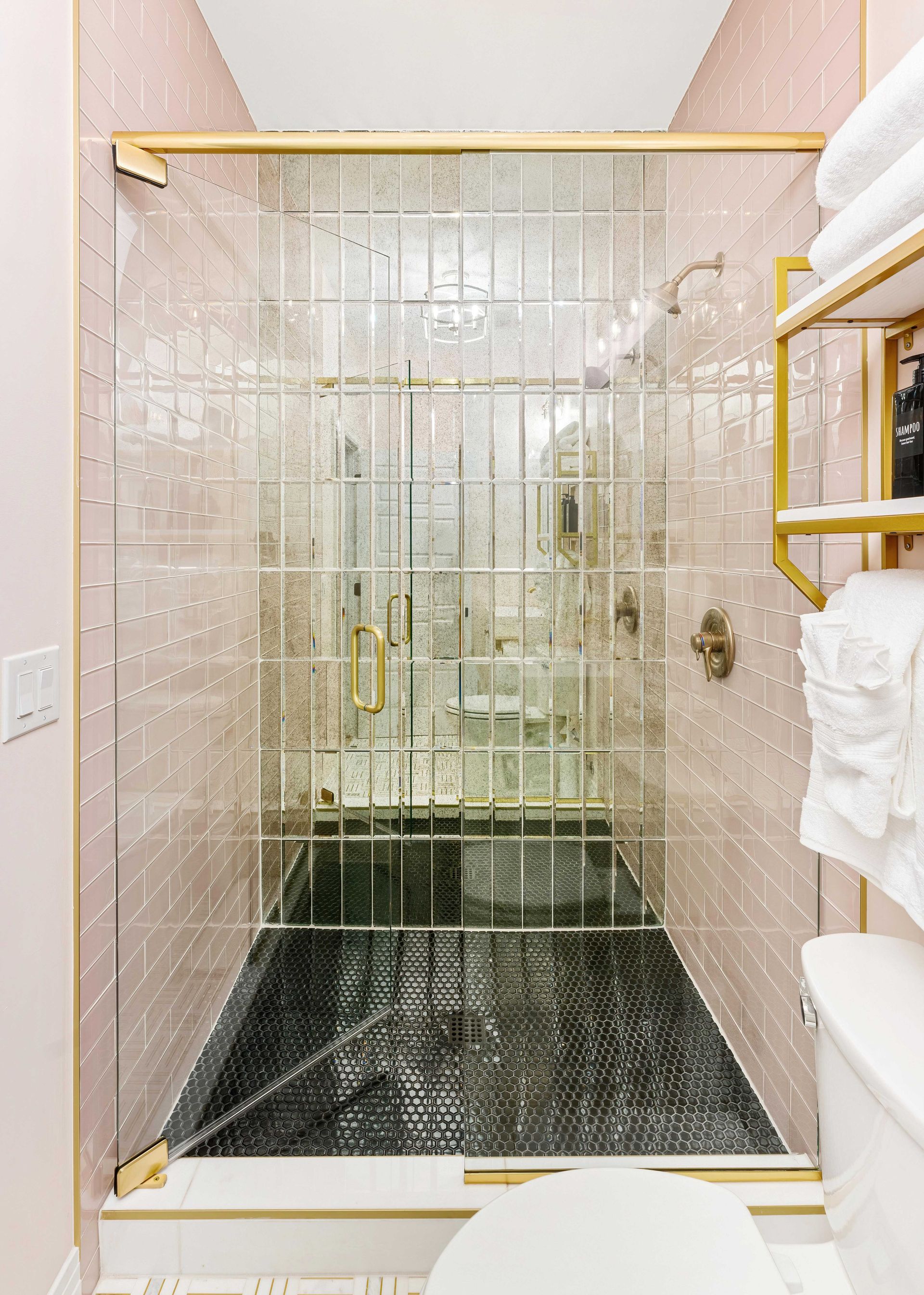 Glass-enclosed shower with gold trim, black tile floor, and white towel in a pink bathroom