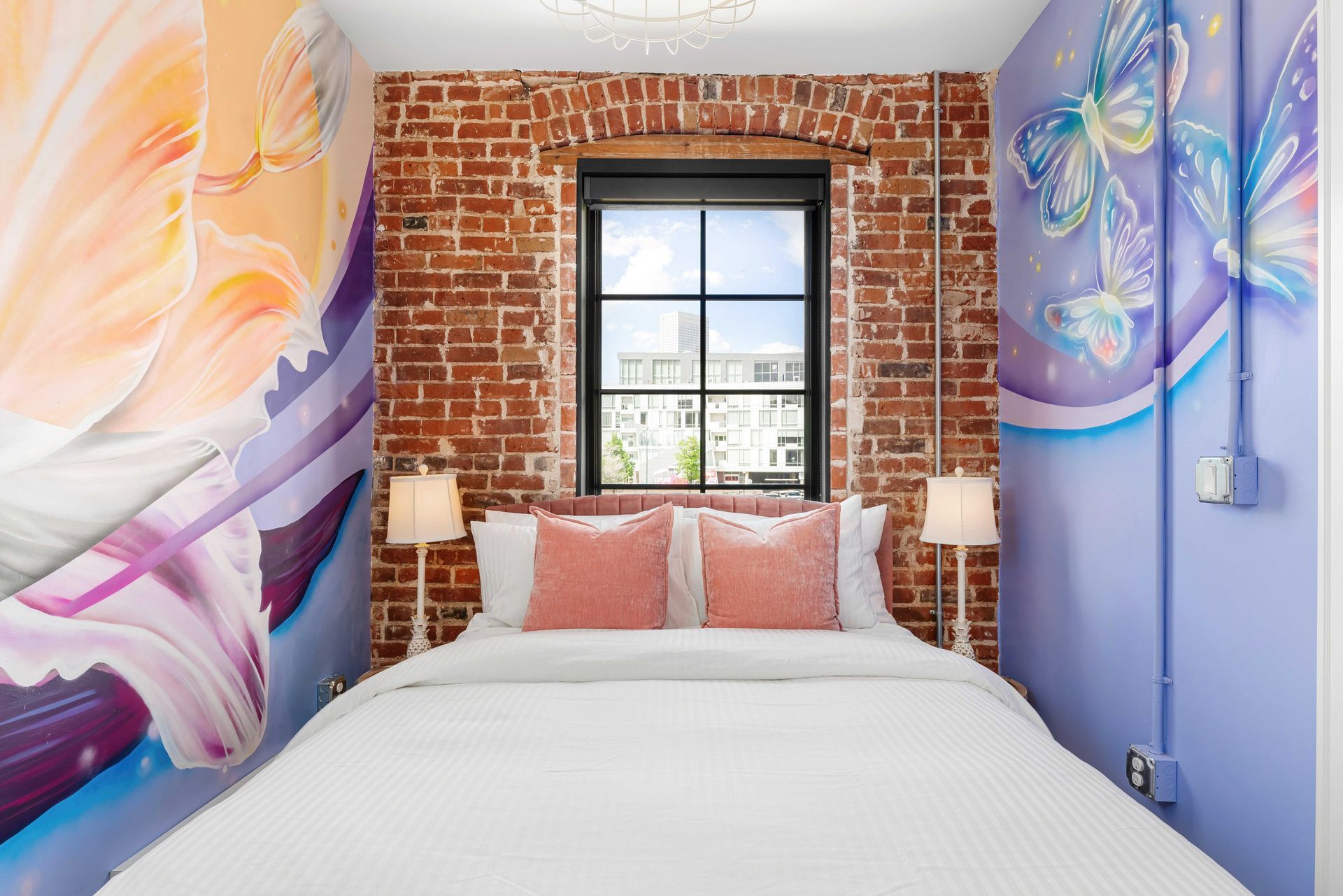 Small bedroom with colorful mural walls, exposed brick, a window, and a neatly made bed with pink pillows