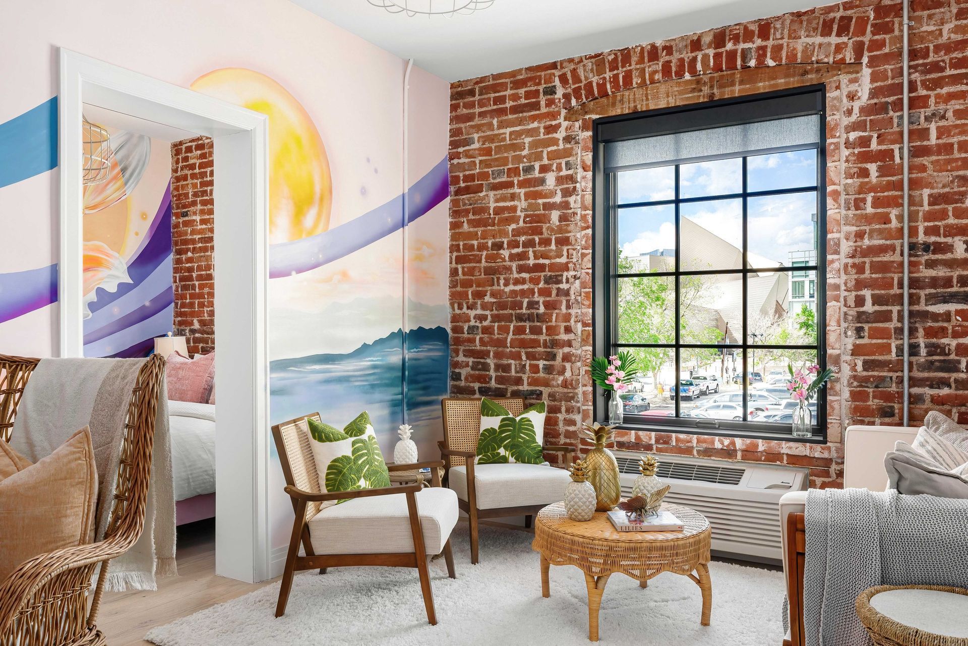 Bright living room with exposed brick, colorful mural, wicker chairs, and a large window.