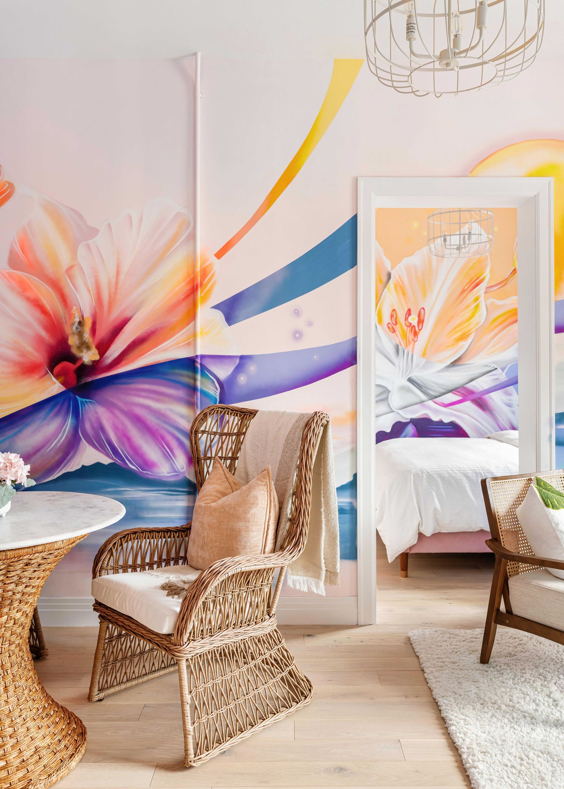 Bright floral mural wall with wicker chairs, gold cushions, and a mirror reflecting a colorful bedroom.