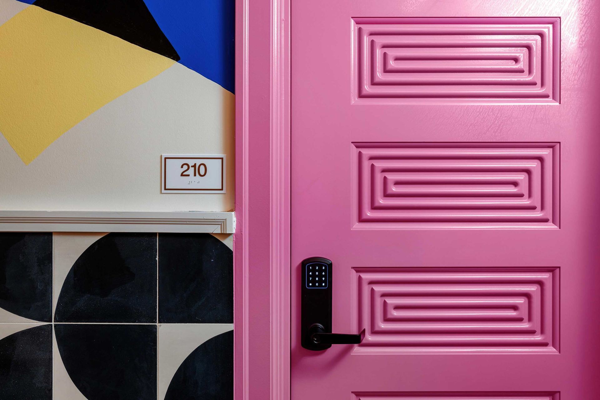 Pink door with keypad lock beside a wall marked 210 and geometric black, yellow, and blue tiles