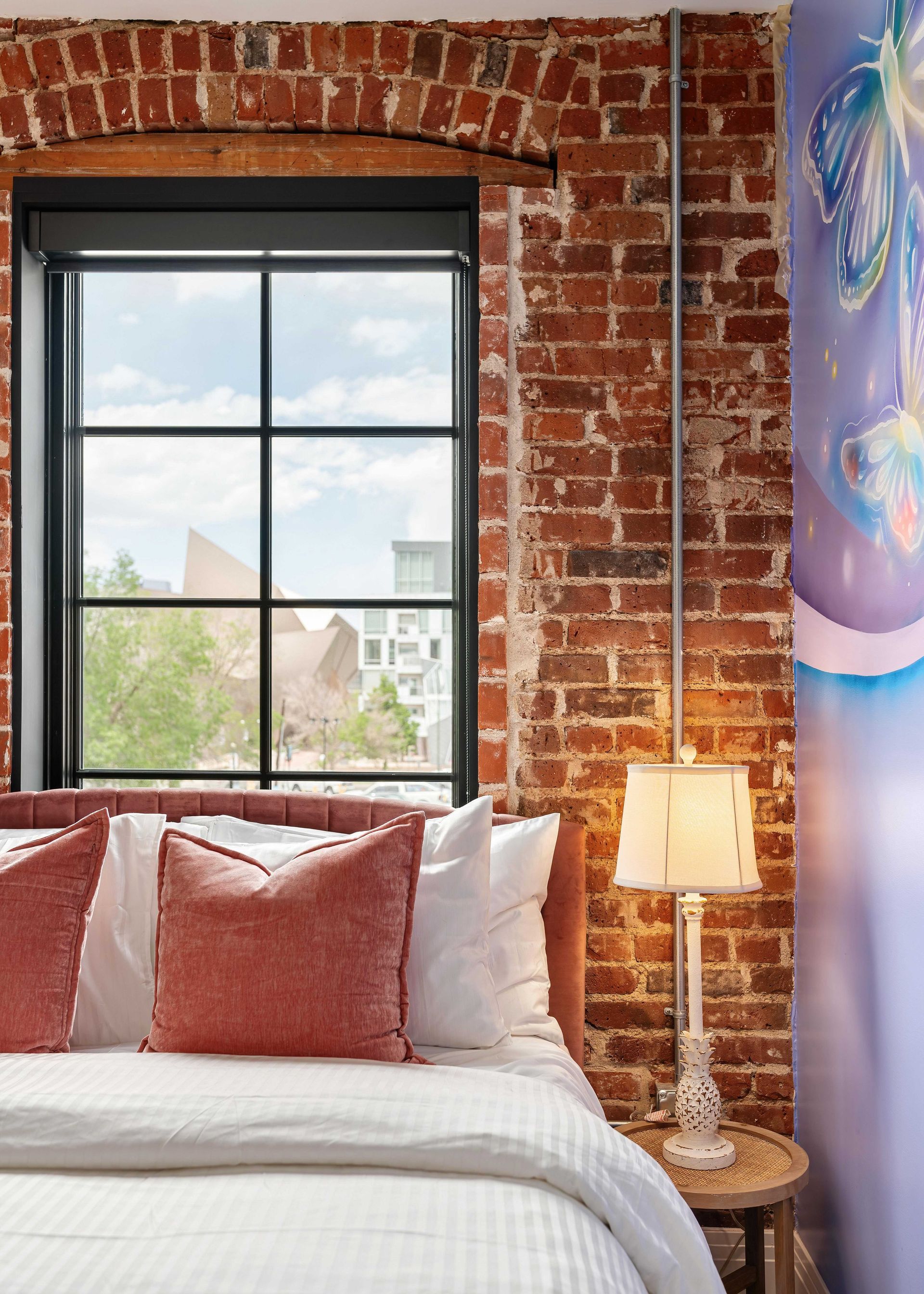 Cozy bedroom with exposed brick wall, black-framed window, white bedding, and a warm bedside lamp.
